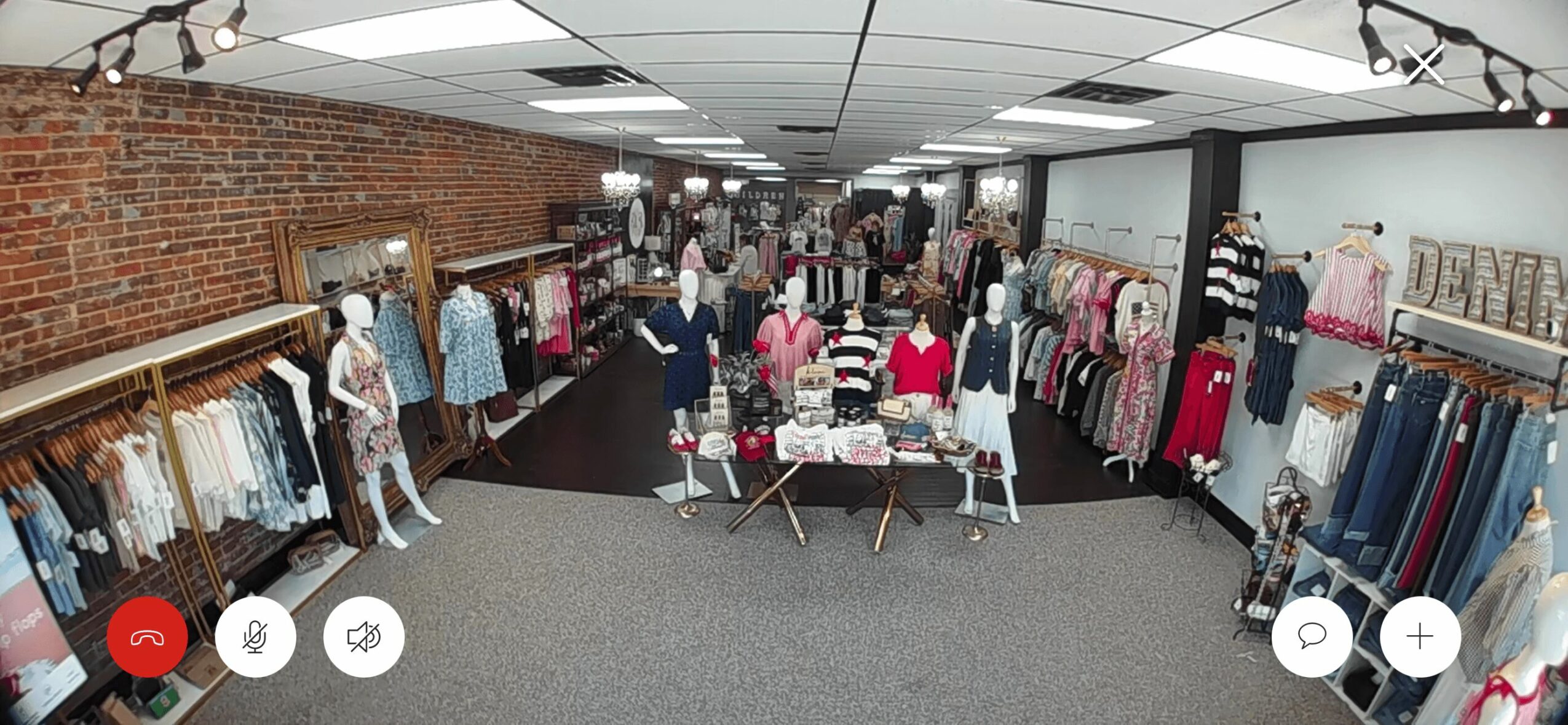 Clothing store interior with racks of clothes, tables, and shoppers browsing, brick wall on the left, ceiling lights overhead.
