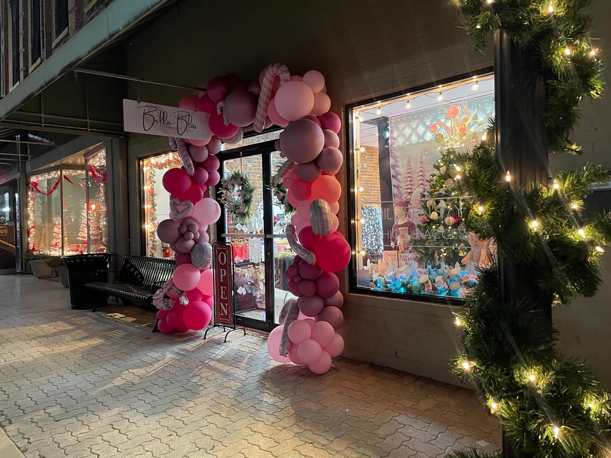 Decorated storefront entrance with pink and white balloon arch, festive lights, and holiday greenery, suggesting a celebration or event.