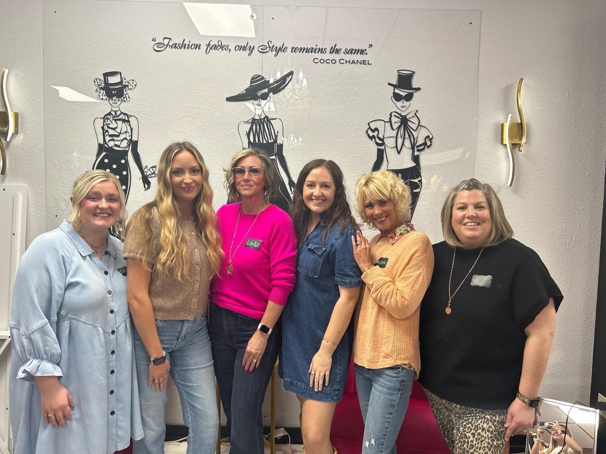 Five women standing together in front of a fashion illustration wall, smiling, in a room with white walls and wall sconces.