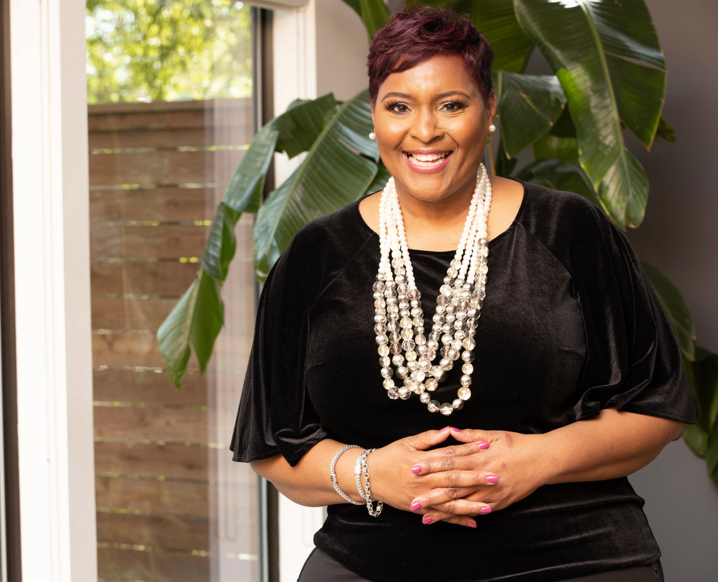 Smiling woman with short hair wearing a black dress and multiple pearl necklaces, standing indoors near large green plants.