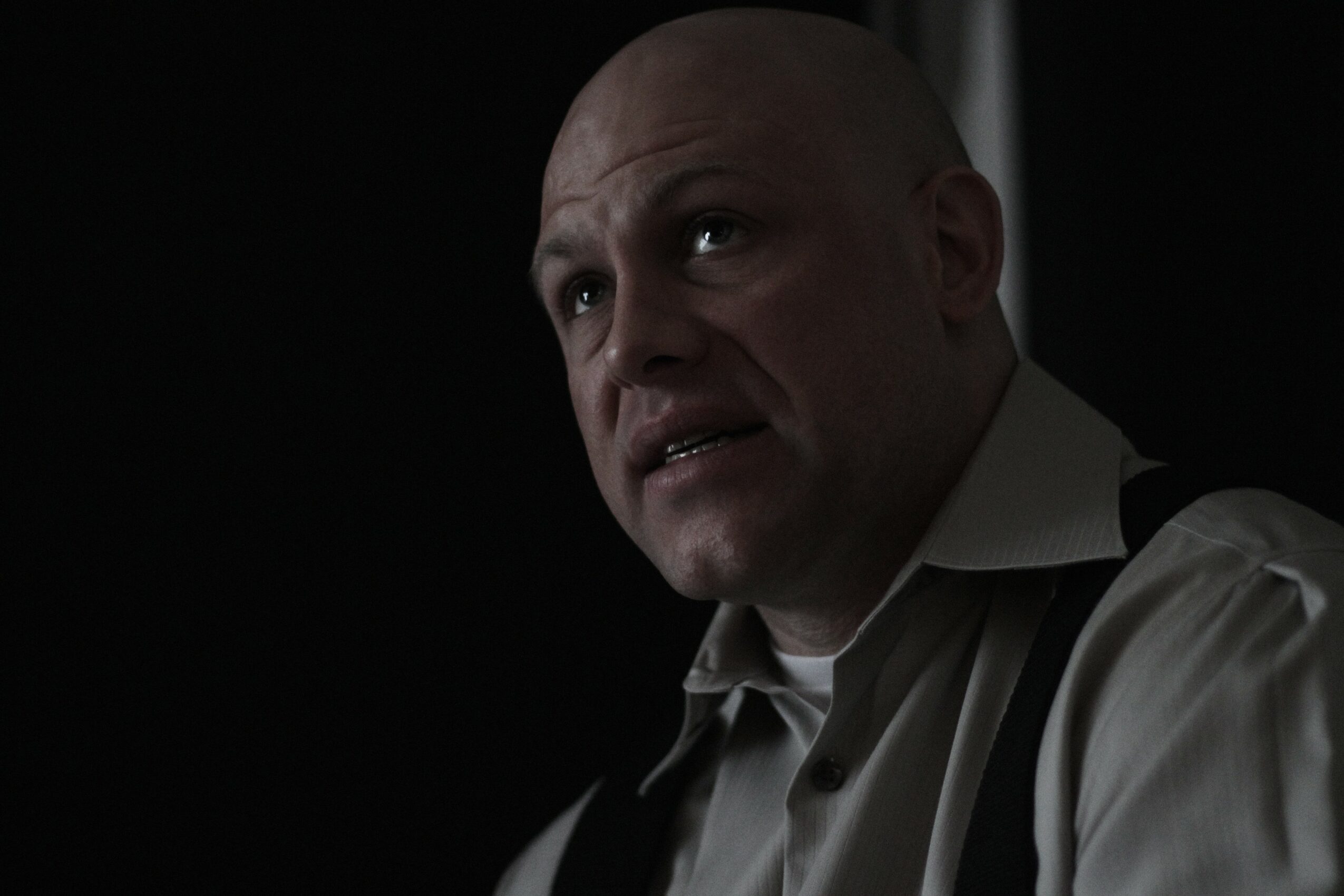 Bald man in a white shirt with suspenders looks to the side in a dark setting.