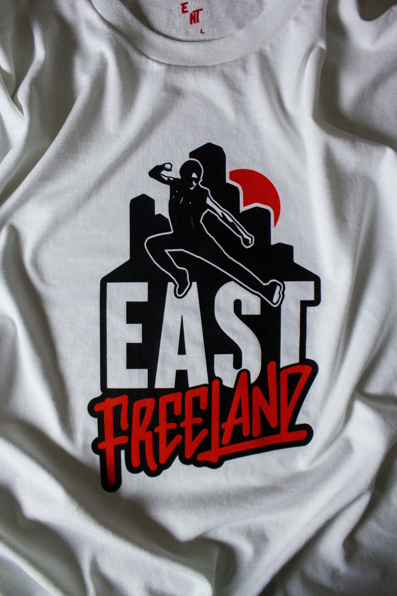 White shirt with a black and red graphic of a woman sitting on a structure, with bold text reading 'EAST FREELAND 12'.