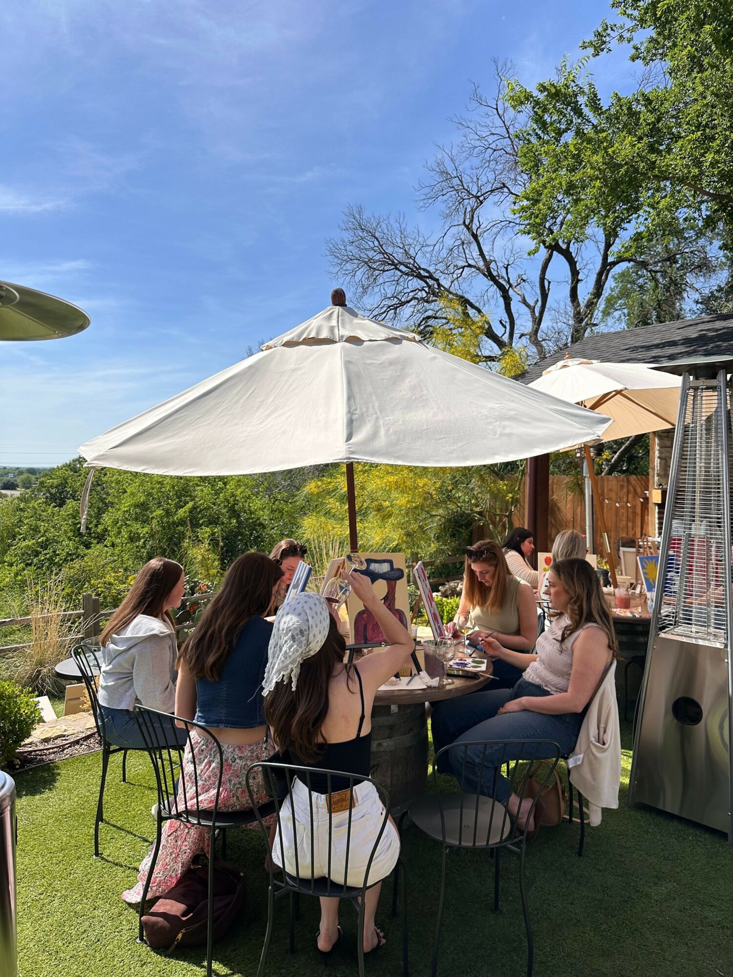 Group of women gathered outdoors under a white umbrella, sitting and standing around a table, with trees and blue sky in background.