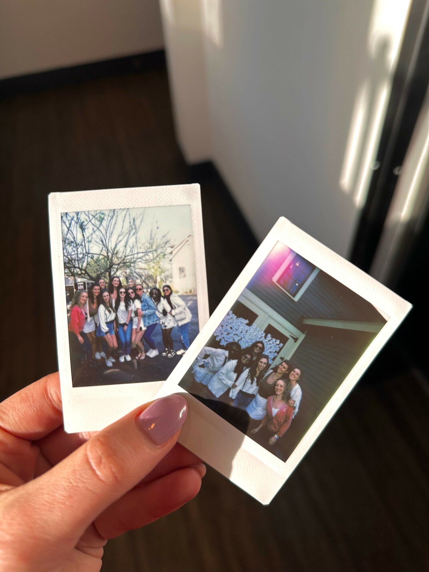 Person holding two printed photos, one outdoors with a group of people, one indoors with a group, sunlight nearby.
