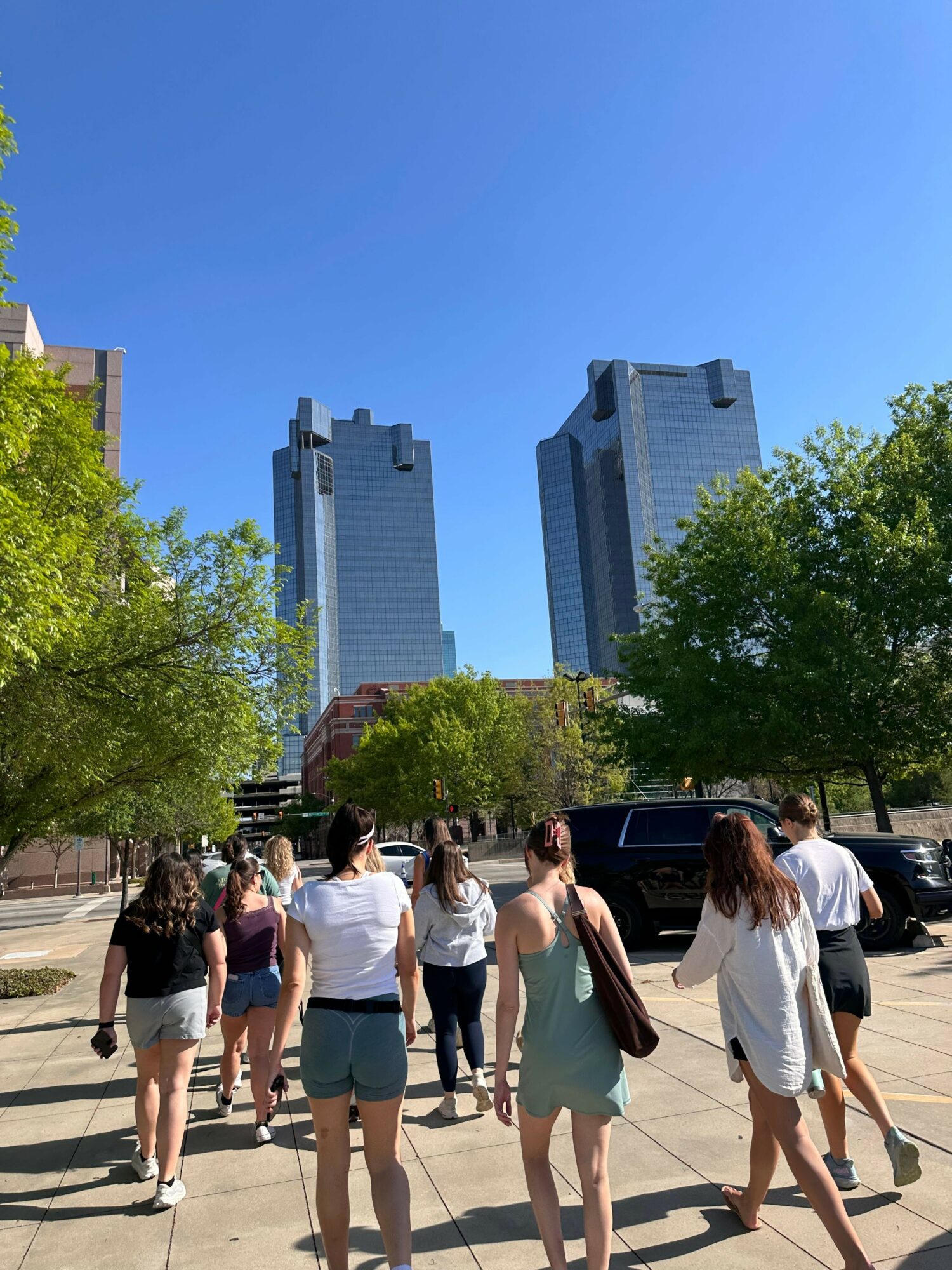 People walking on a city sidewalk with tall buildings and trees, under a clear blue sky.