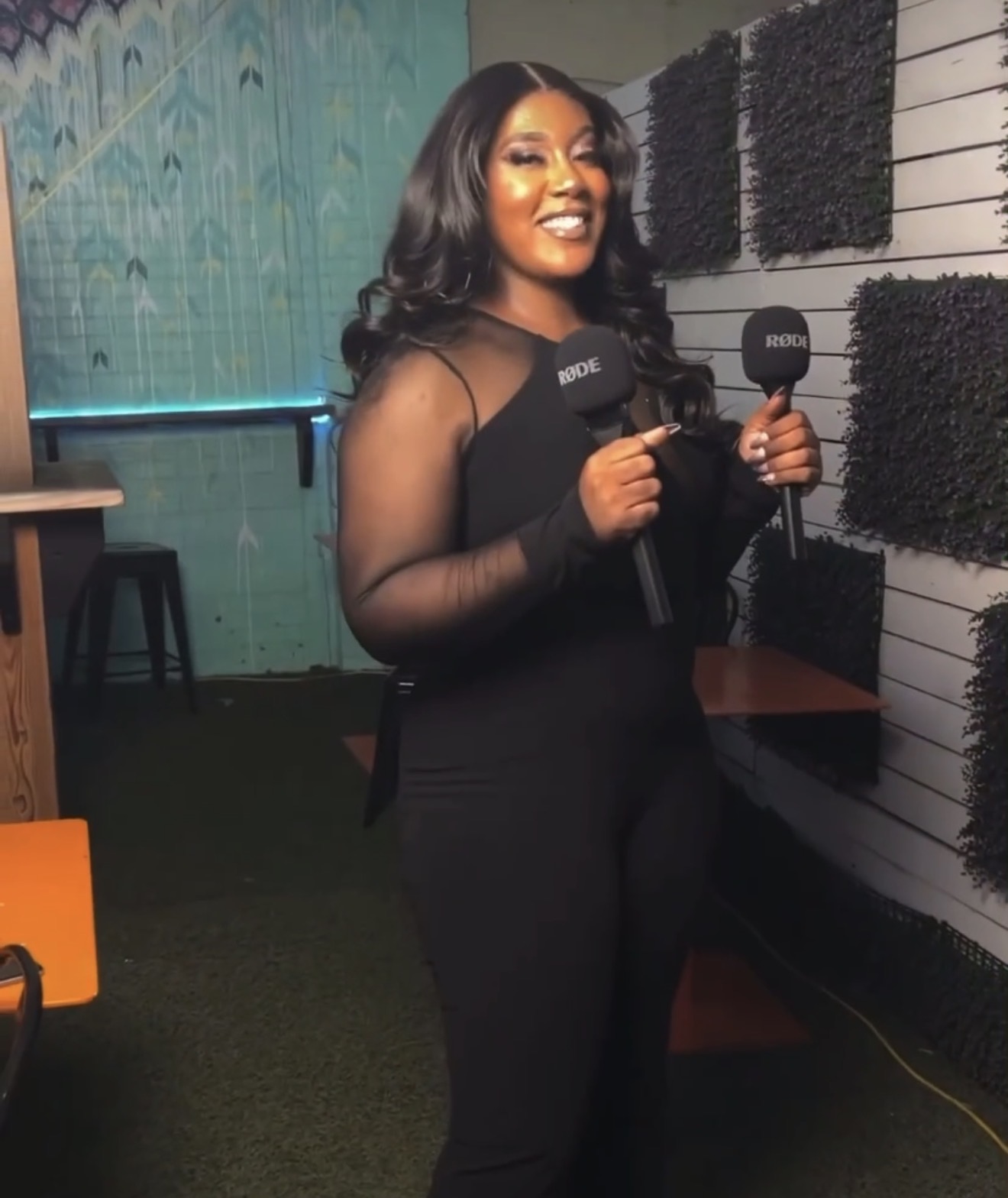 Woman holding two microphones, smiling, standing indoors with textured wall and furniture behind her.