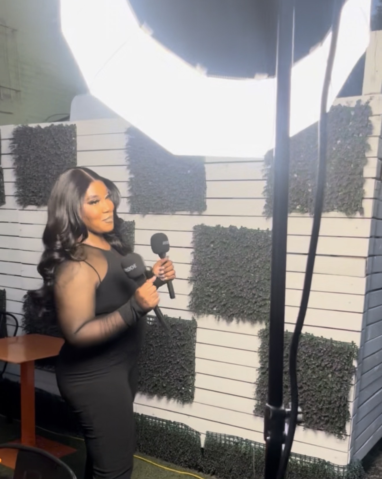 Woman in black dress holding a microphone, standing indoors with a white wall and decorative black panels behind her.