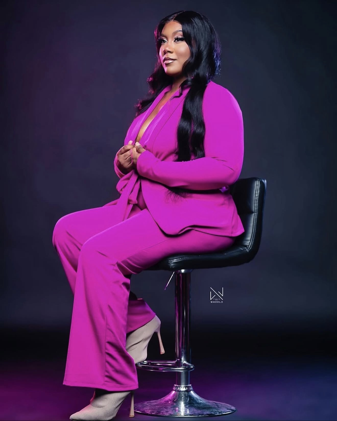 Woman in bright pink suit sitting on a black chair against a dark background, smiling, with long black hair.