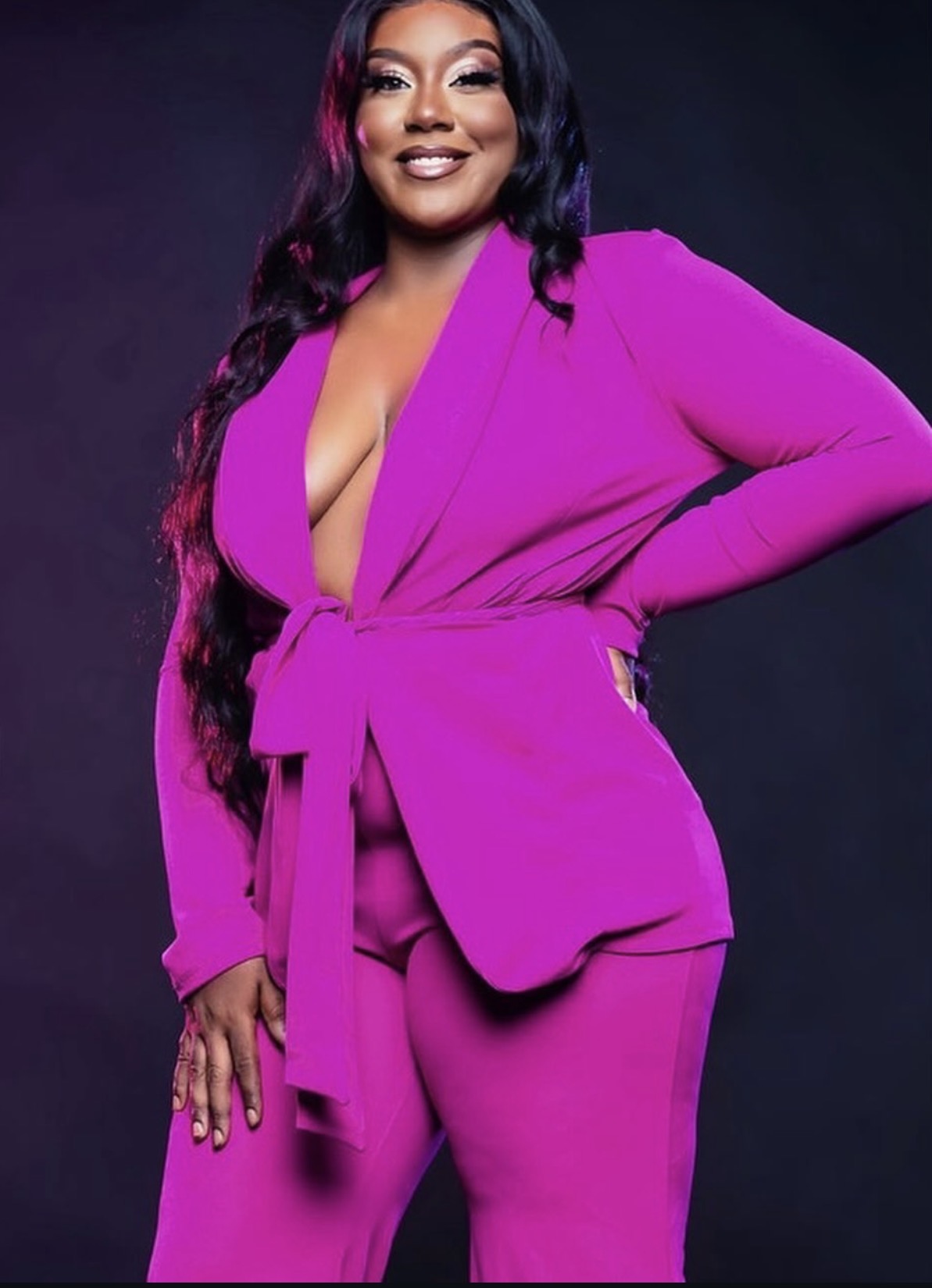 Woman in a bright pink suit with a deep neckline, smiling, with long dark hair, standing against a dark background.