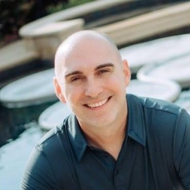 Smiling man with a shaved head wearing a dark shirt outdoors near boats or water.