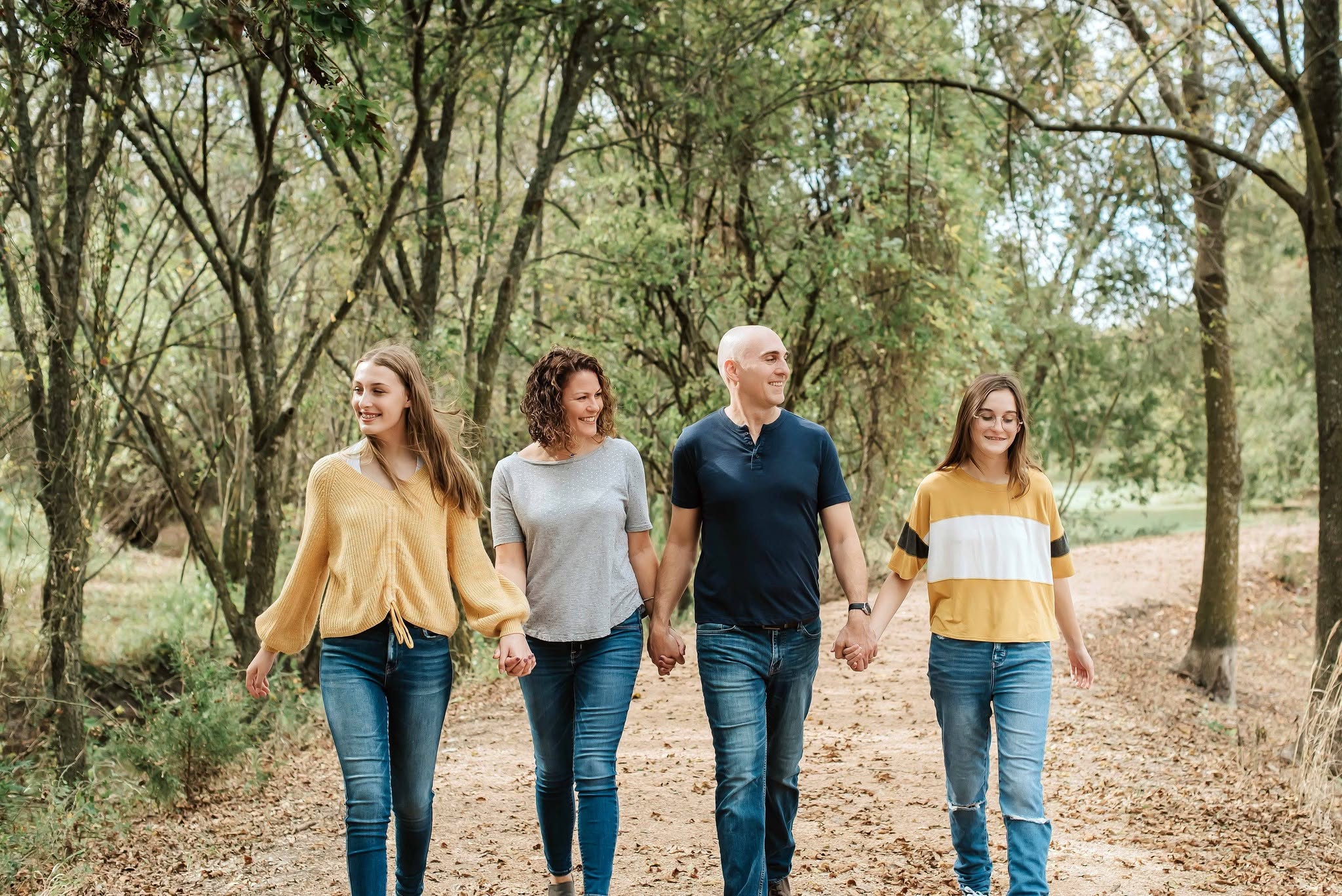 Four people walk hand in hand through a wooded park, smiling and enjoying the outdoors.