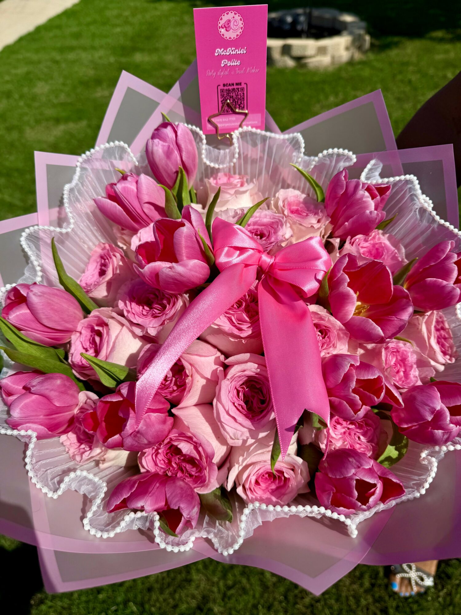 Bouquet of pink tulips and roses wrapped in pink and purple paper with a pink ribbon, outdoors on grass.