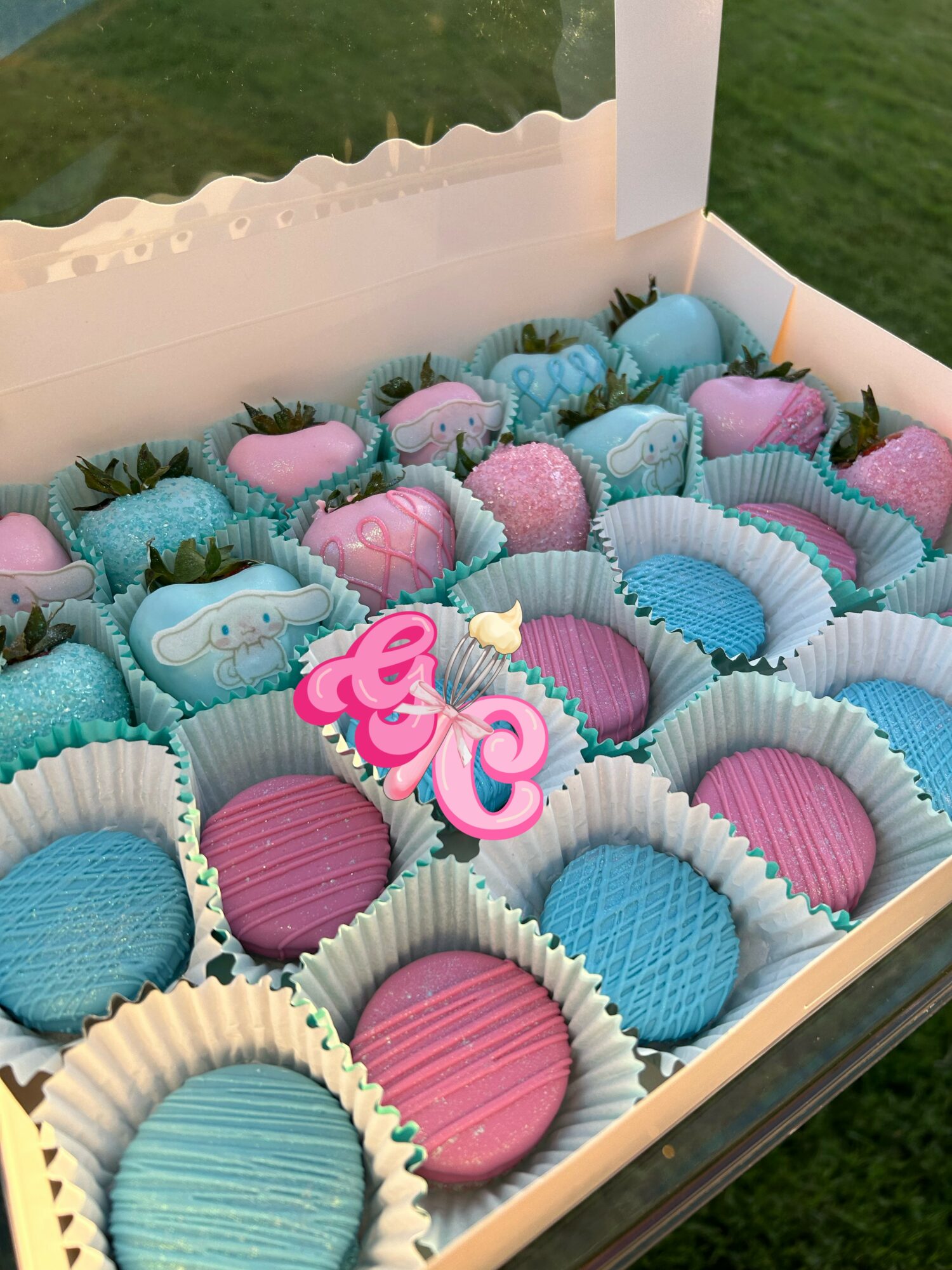 Box of pink and blue decorated cookies with strawberries, some with pink and blue icing, on a grassy background.
