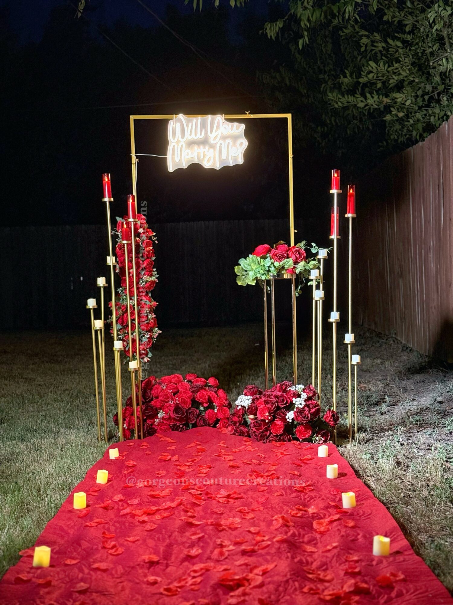 Night scene with floral decorations, candles, and a neon sign reading 'Will You Marry Me' over a red carpet pathway.