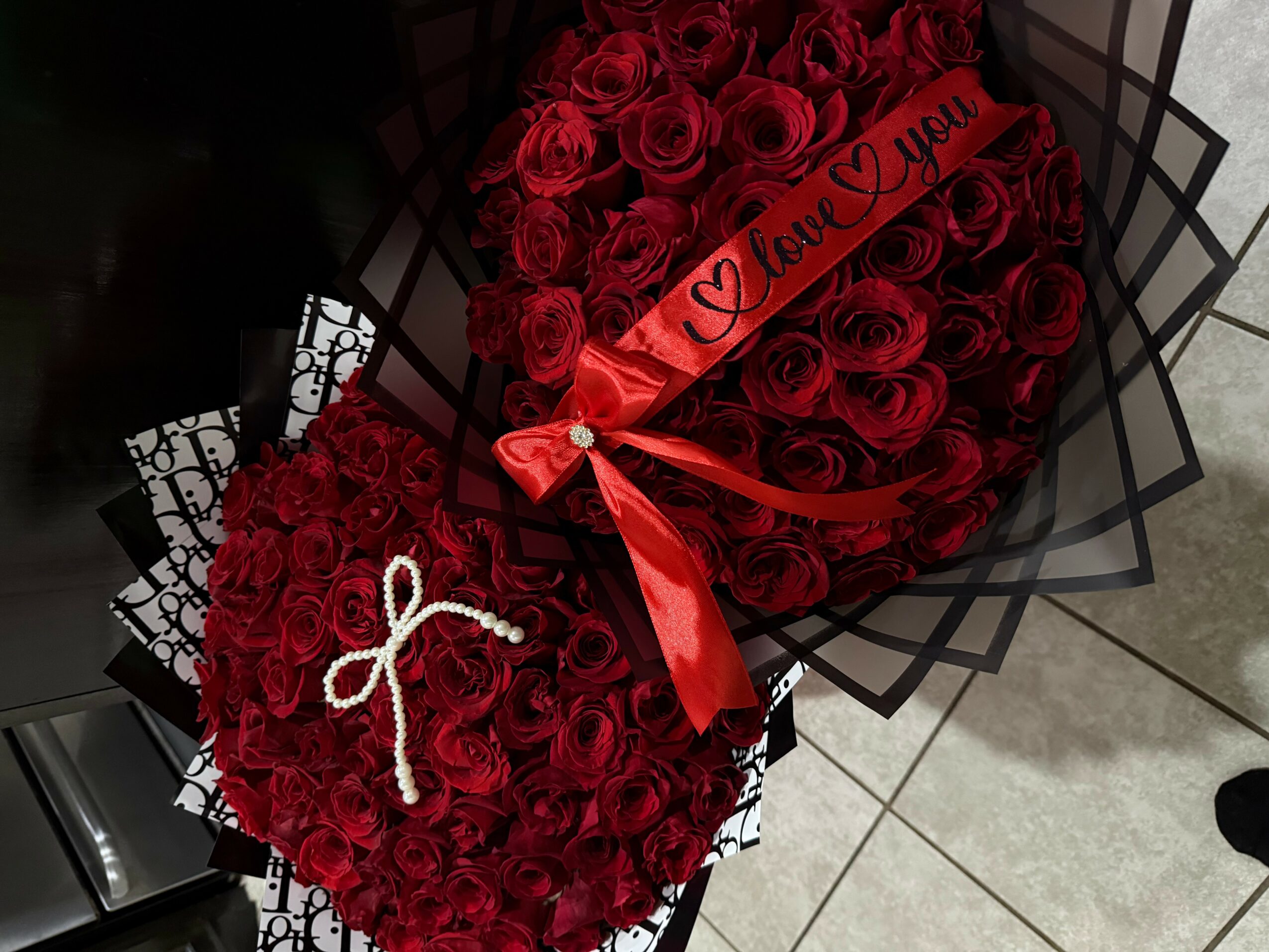 Two heart-shaped bouquets of red roses, one with a red ribbon and a message, the other with a white bow, on tiled floor.