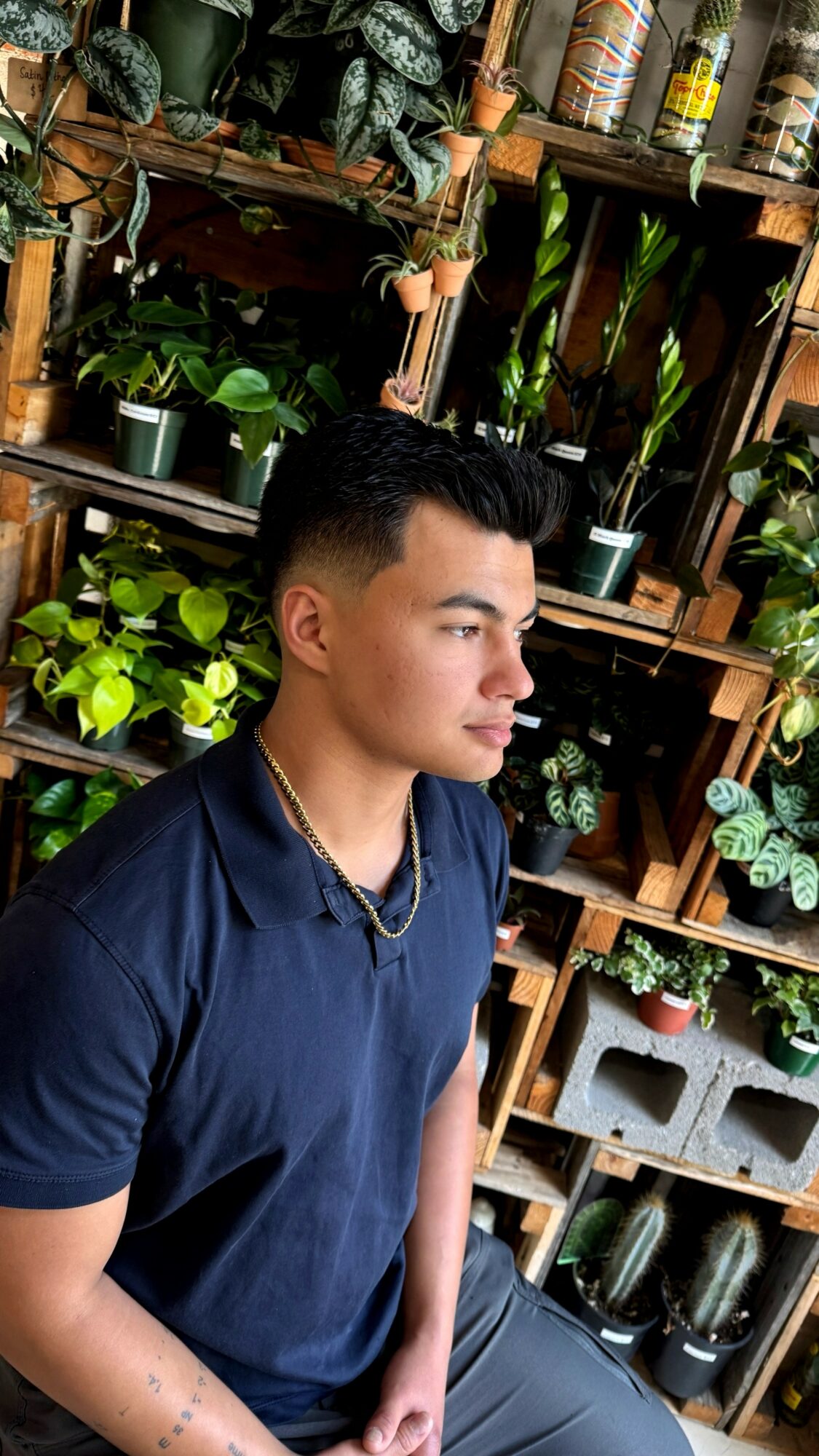 Young man with short dark hair wearing a navy shirt, standing in front of a wooden plant shelf with various potted plants.