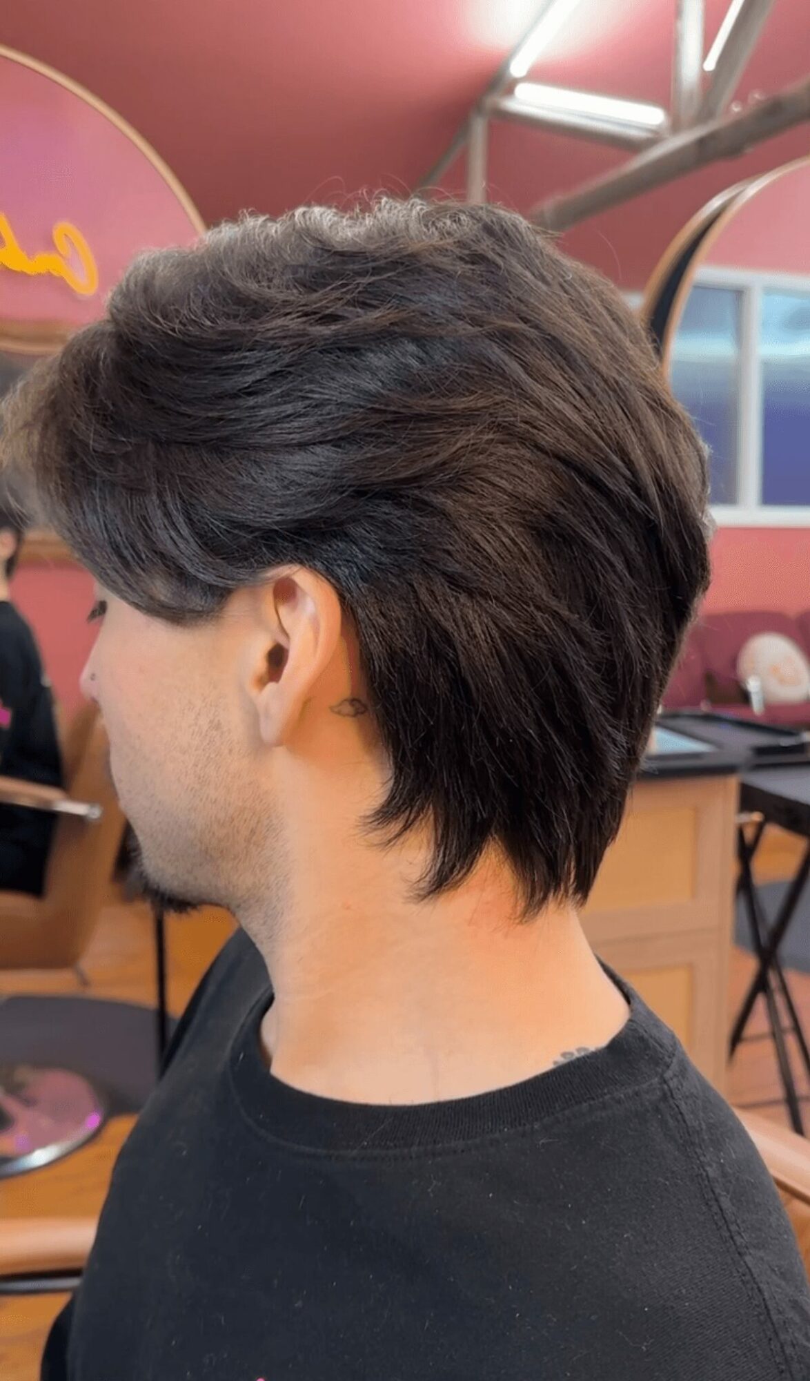 Side view of a person with a layered, dark hairstyle in an indoor setting with pink and purple decor.