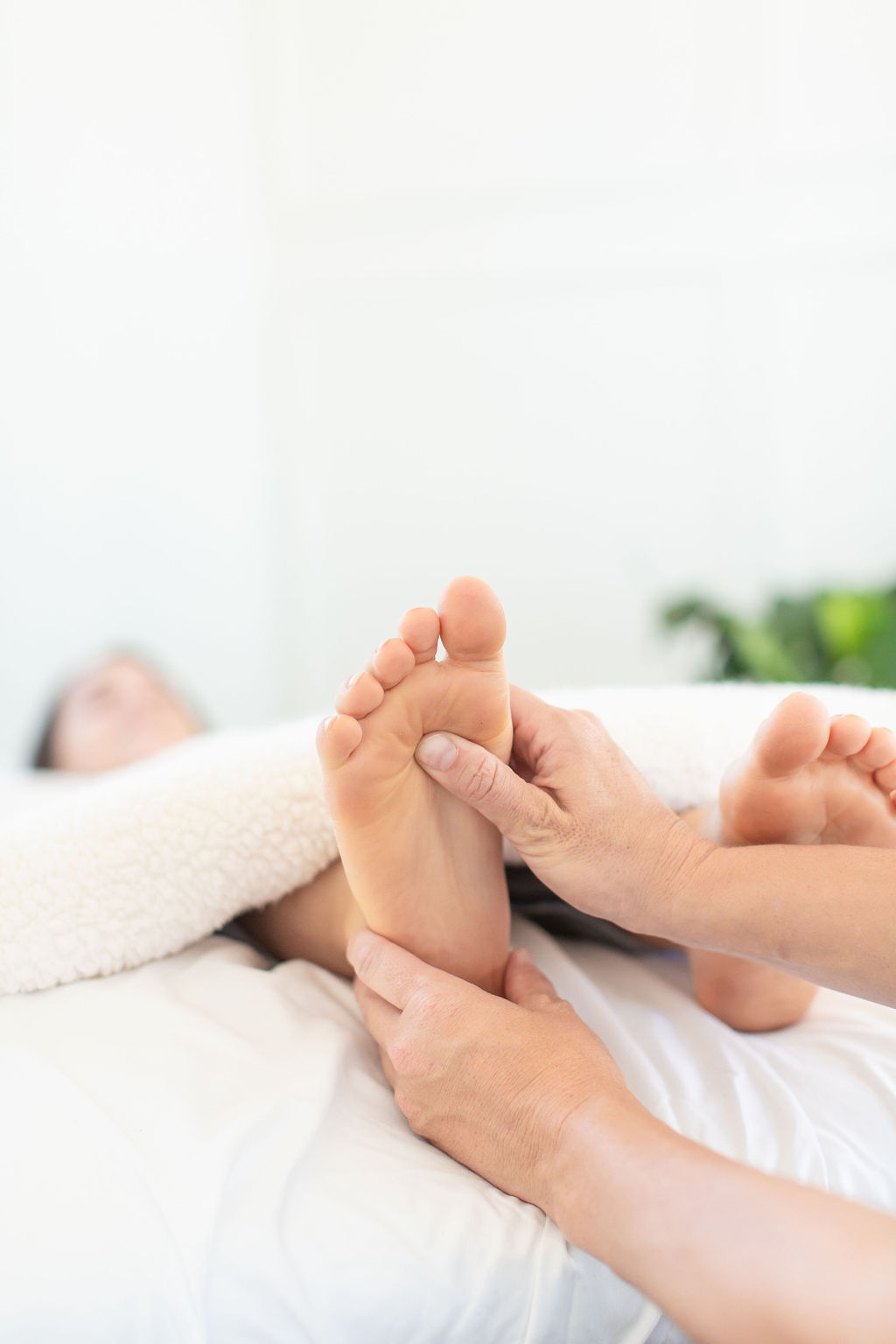 Person lying in bed receiving foot massage, with a blurred background and a plant. 20 words.