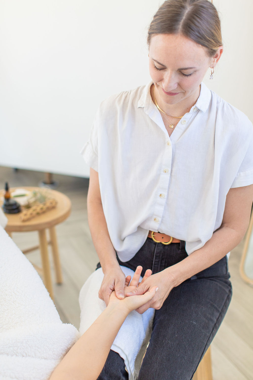 Woman with brown hair in a ponytail, wearing a white shirt, holding a person's hand gently, in a bright room.