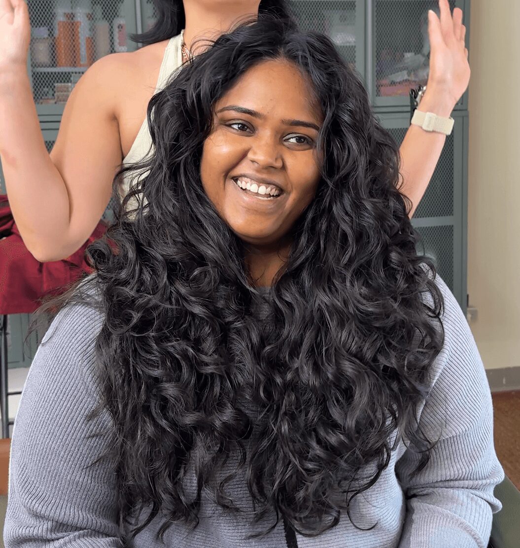 Smiling woman with long, curly hair wearing a gray sweater, in a room with cabinets in the background.