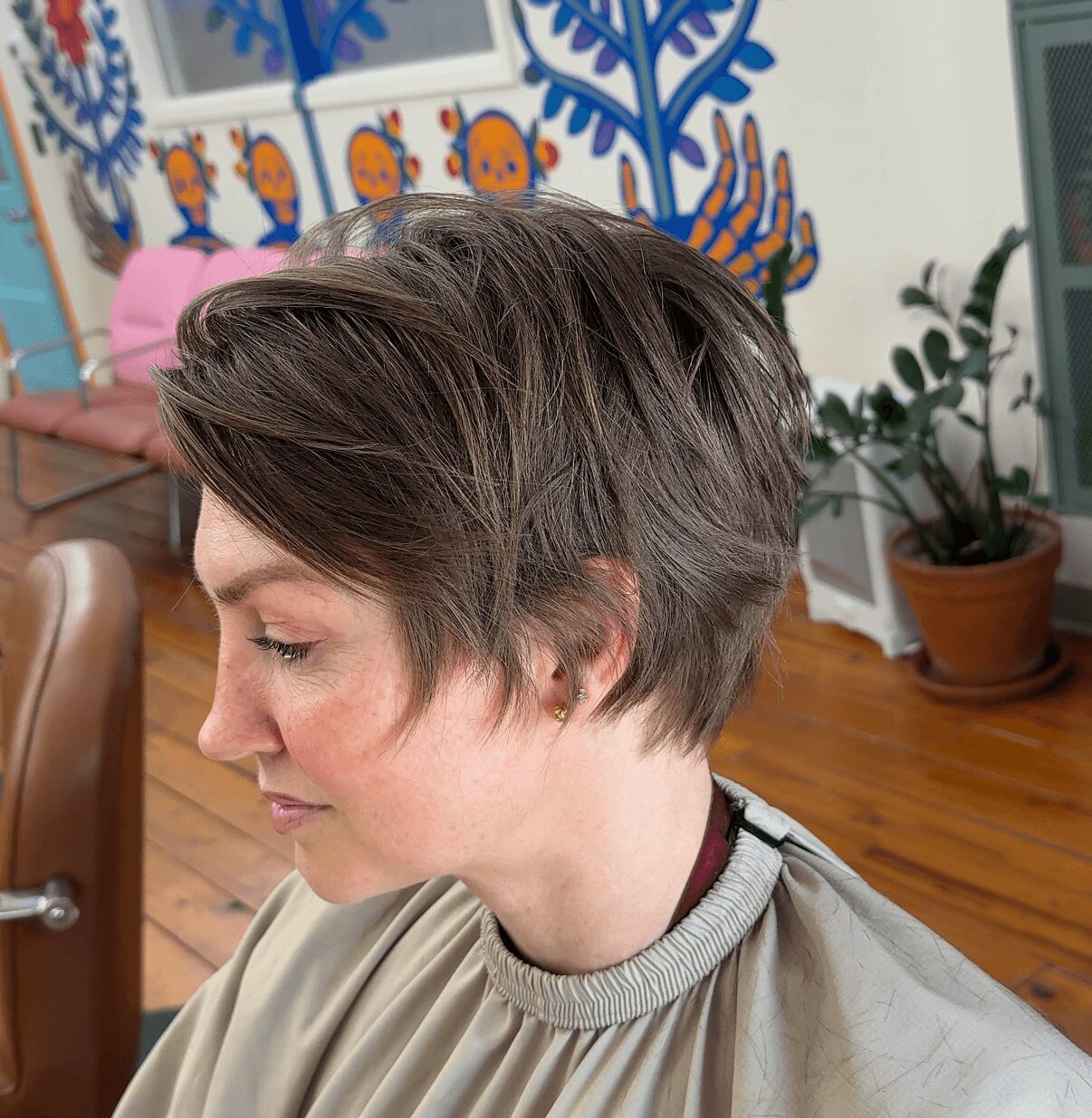 Side profile of a woman with short, layered hair in a room with wooden flooring and a colorful mural.