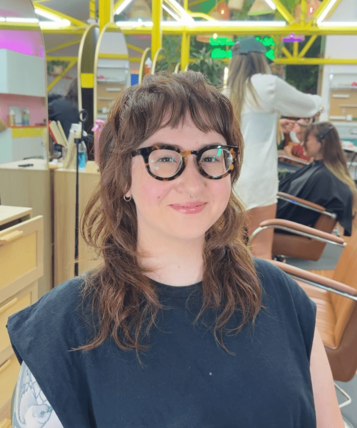 Young woman with wavy brown hair and glasses smiling in a colorful indoor setting.