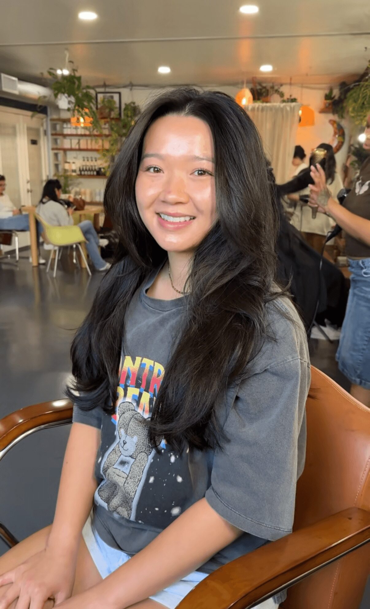 Young woman with long dark hair smiling, sitting in a restaurant or cafe, wearing a gray graphic t-shirt.