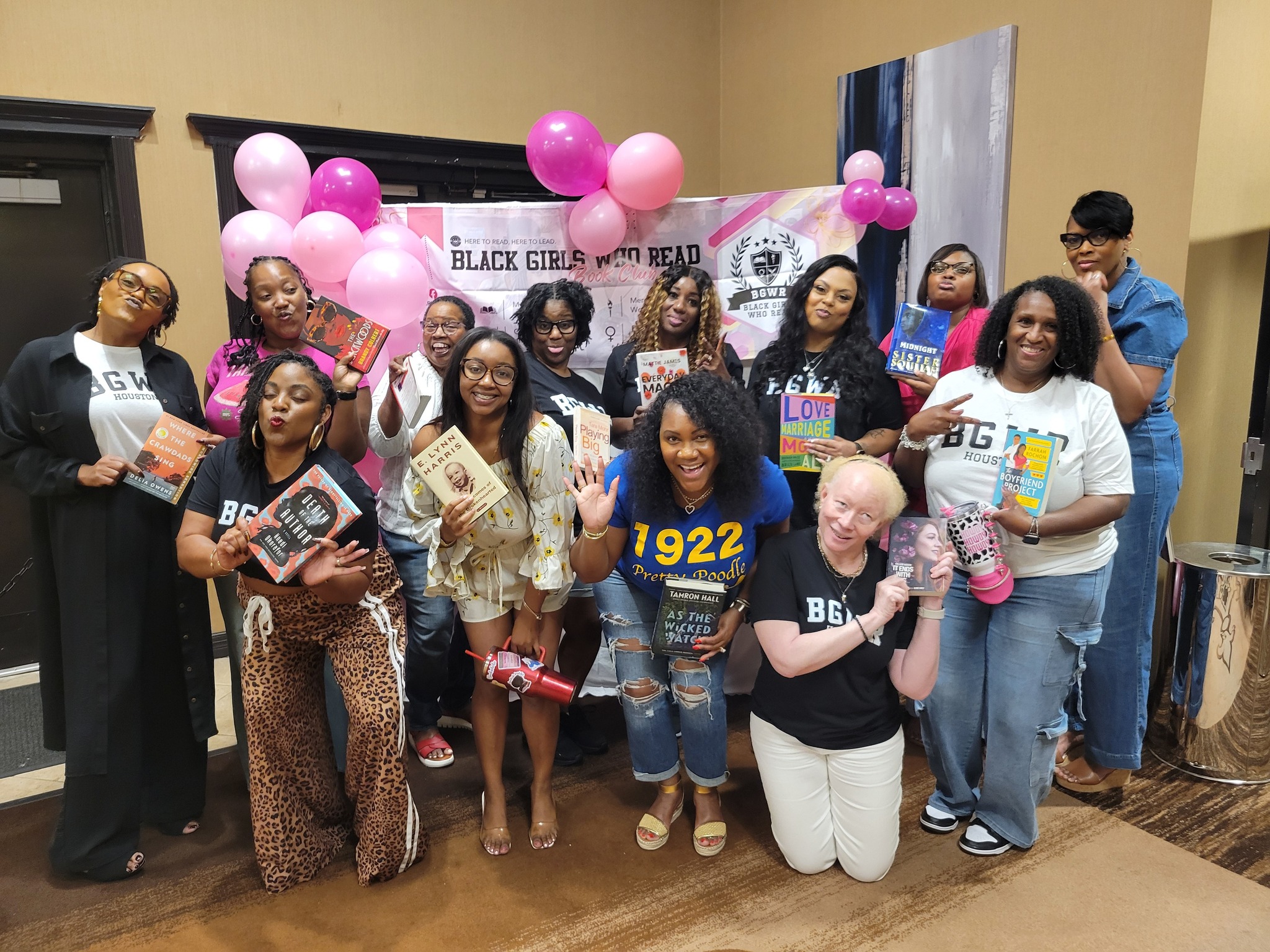 Group of diverse women and girls holding books and pink balloons at a Black Girls Read event, with a banner in the background.