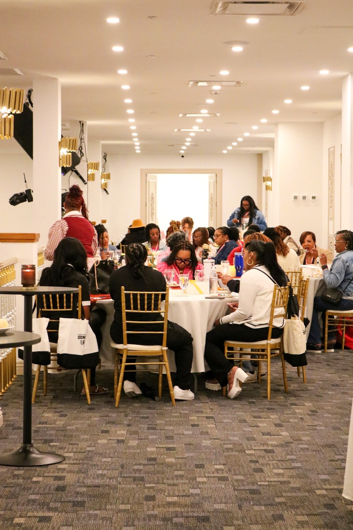 People seated at round tables in a well-lit banquet hall, engaged in conversation, with a white curtain backdrop.