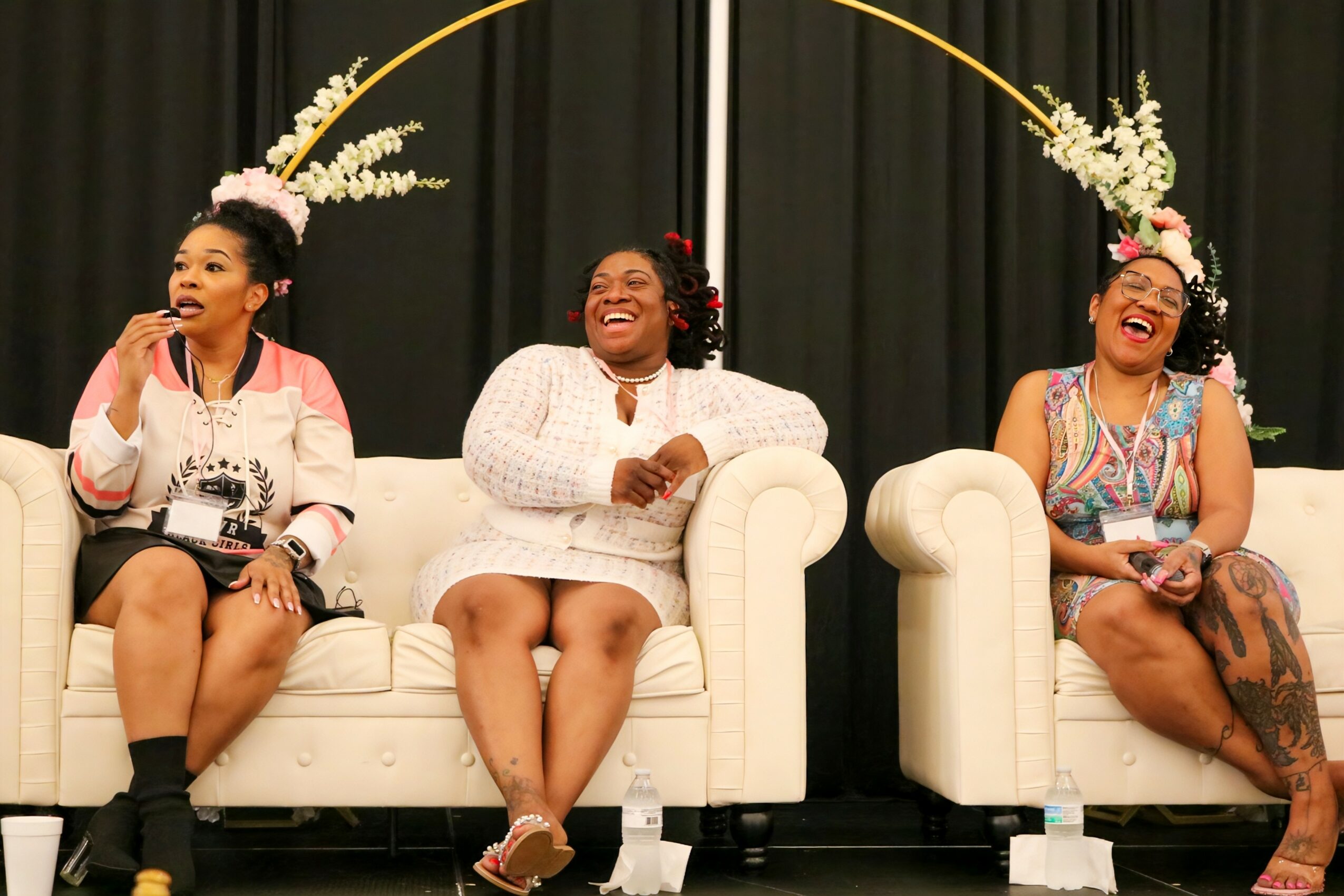 Three women sitting on white sofas, smiling and laughing, with floral decorations above them, black curtains in the background.