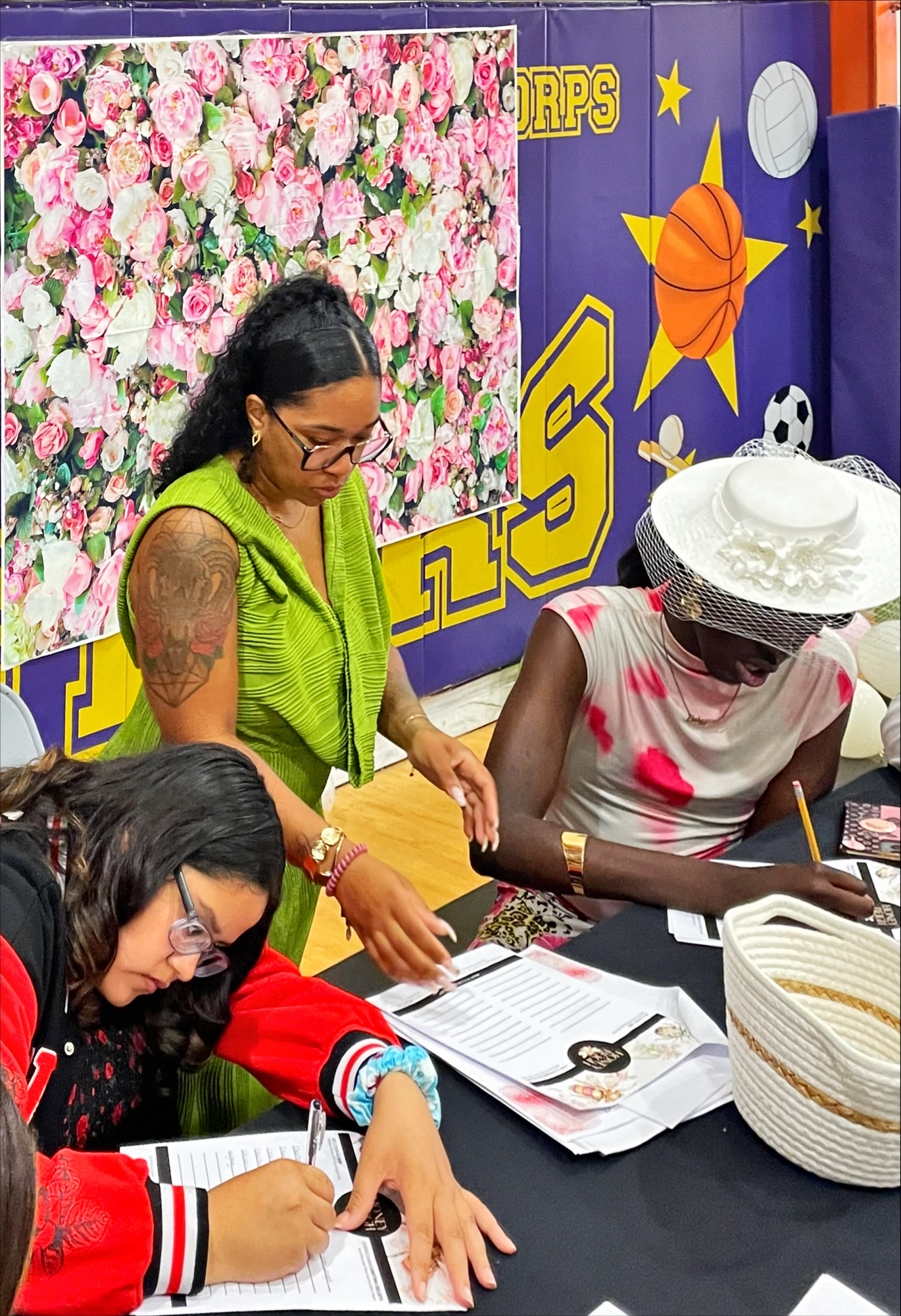 Two women sitting at a table, one in a green top and glasses, the other in a white hat and pink dress, with a floral background.