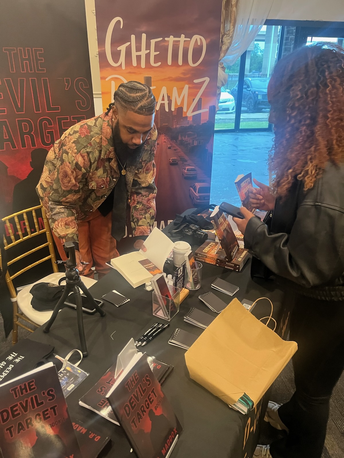 Two people at a table with books and promotional materials, one person with braided hair and the other with curly hair, in front of posters. Word 'Ghetto' visible.