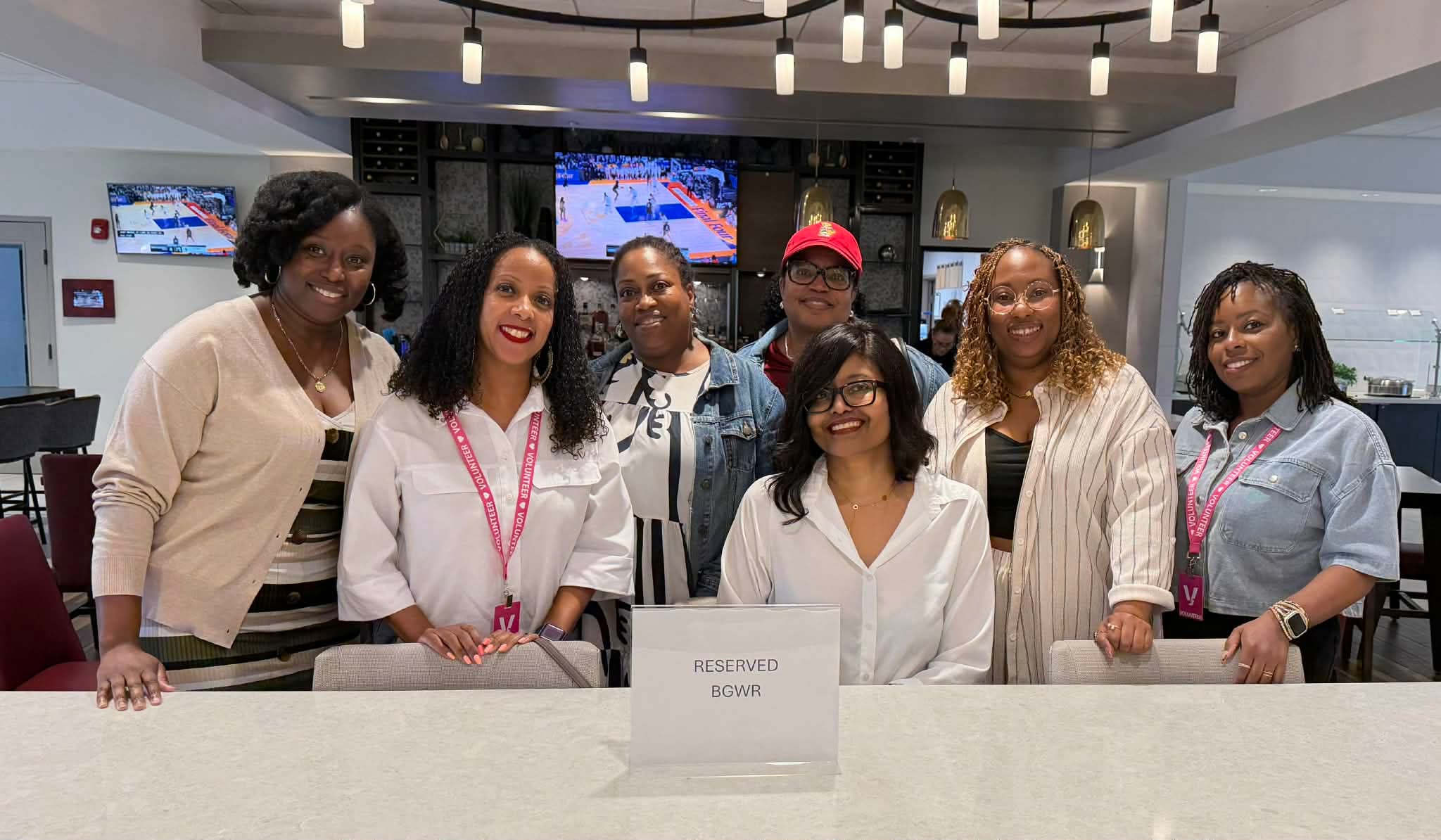 Group of seven women standing behind a reception desk in a modern office, smiling, with screens and lighting fixtures in the background.