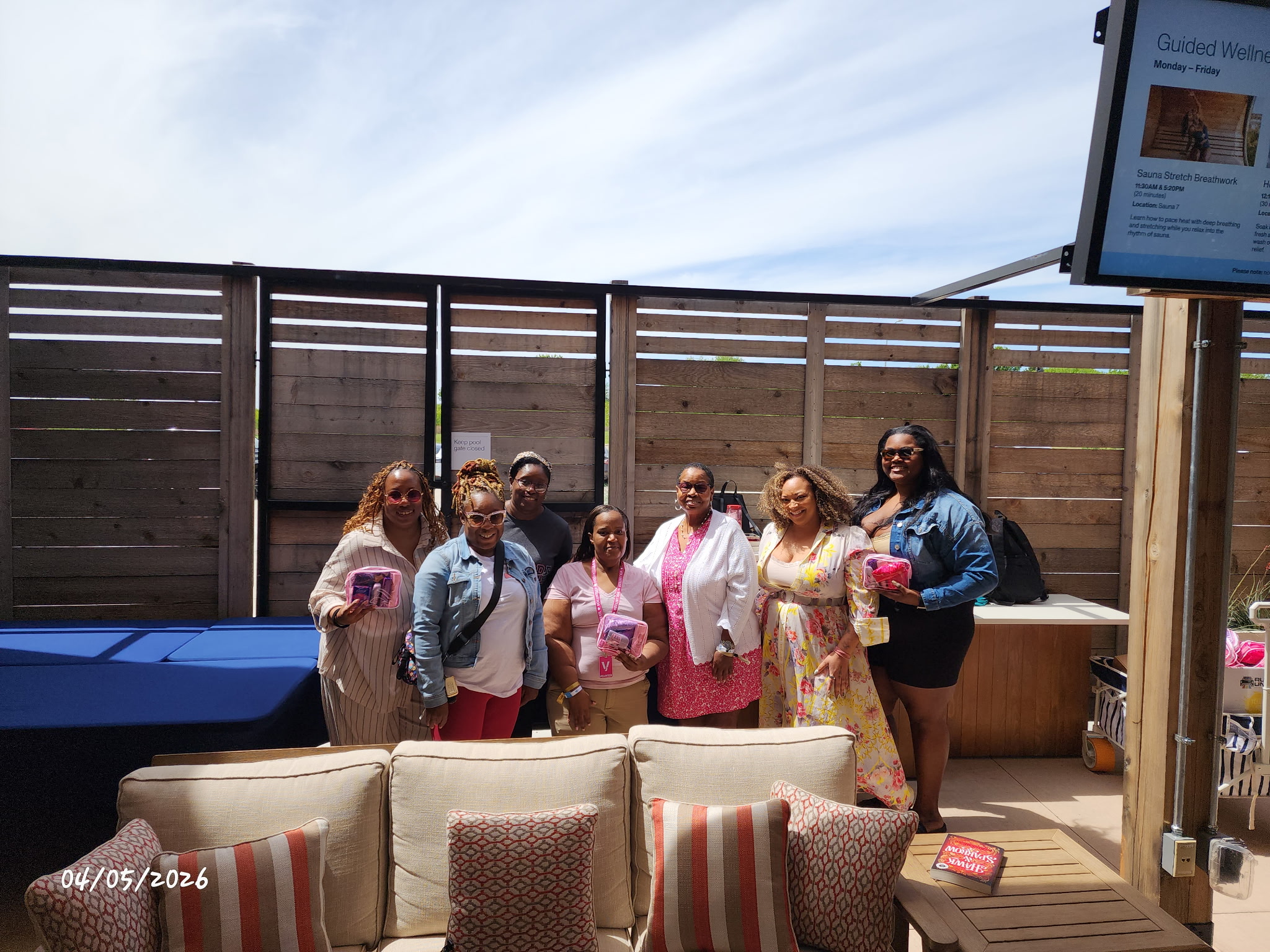 Group of seven women standing outdoors near a wooden fence, some holding drinks, with a blue sky overhead.