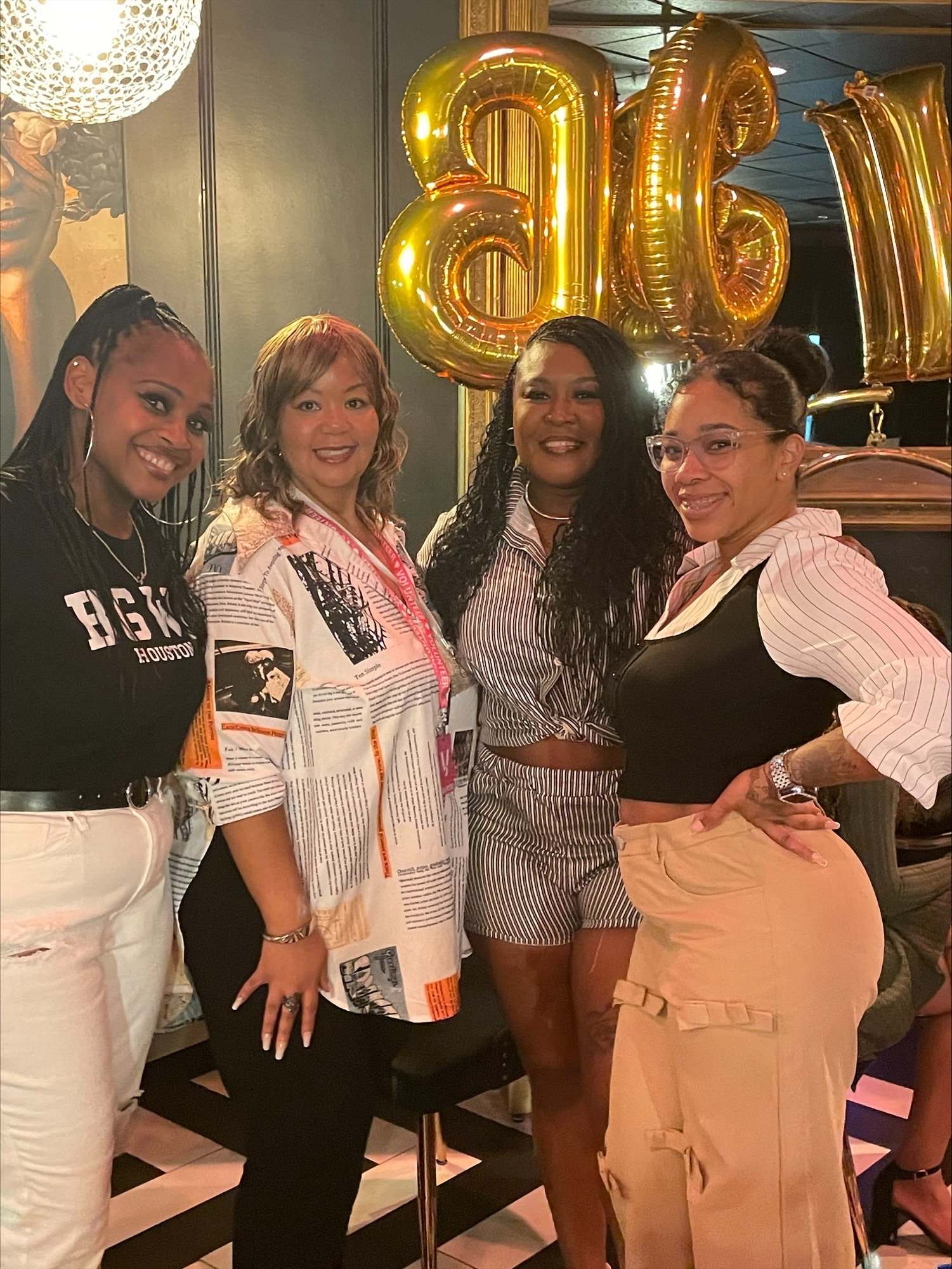 Four women standing together indoors, celebrating with gold balloons, smiling, and dressed casually.
