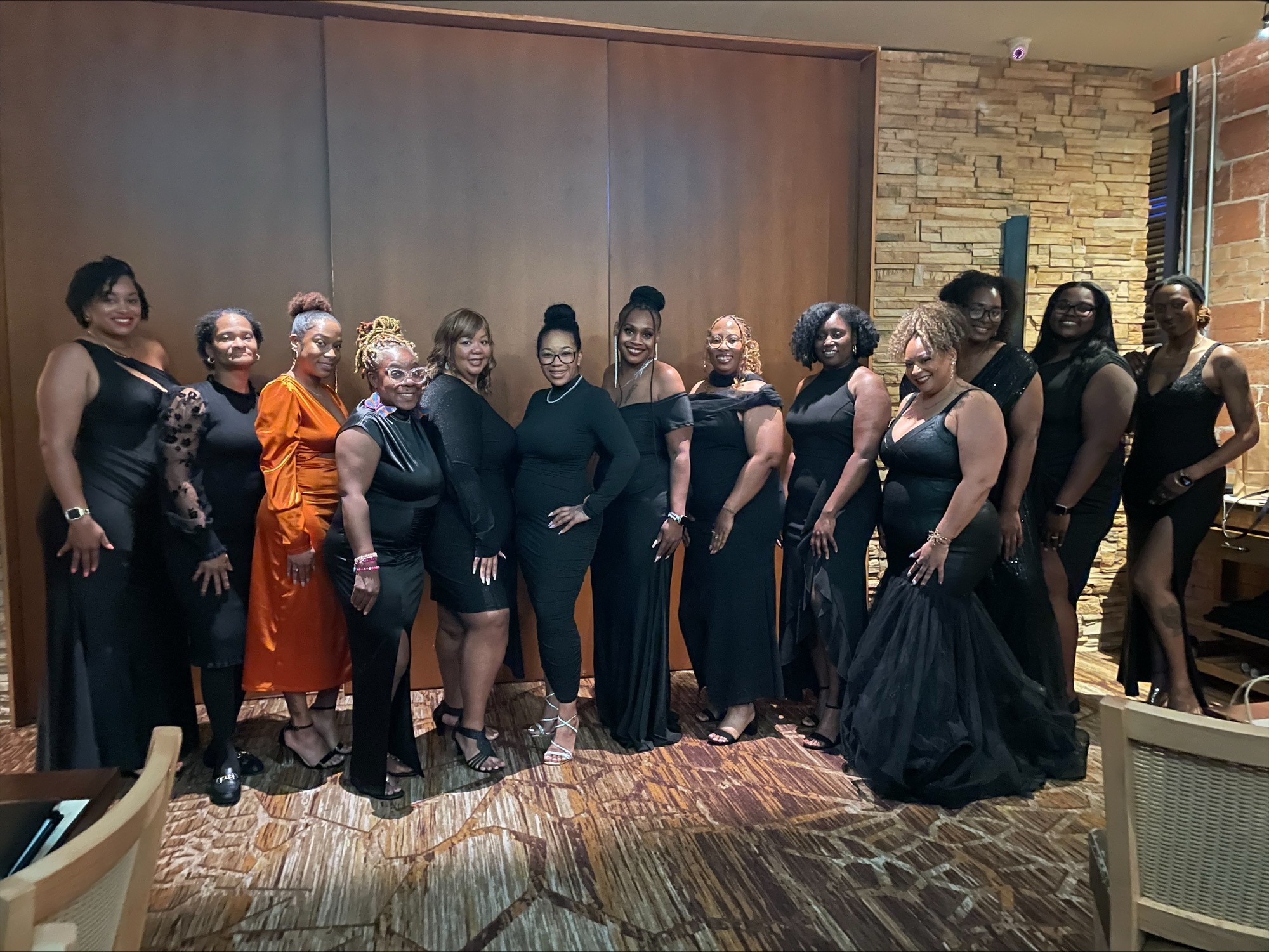 Group of women in black dresses standing indoors, some smiling, with a brick wall and wooden panel background.