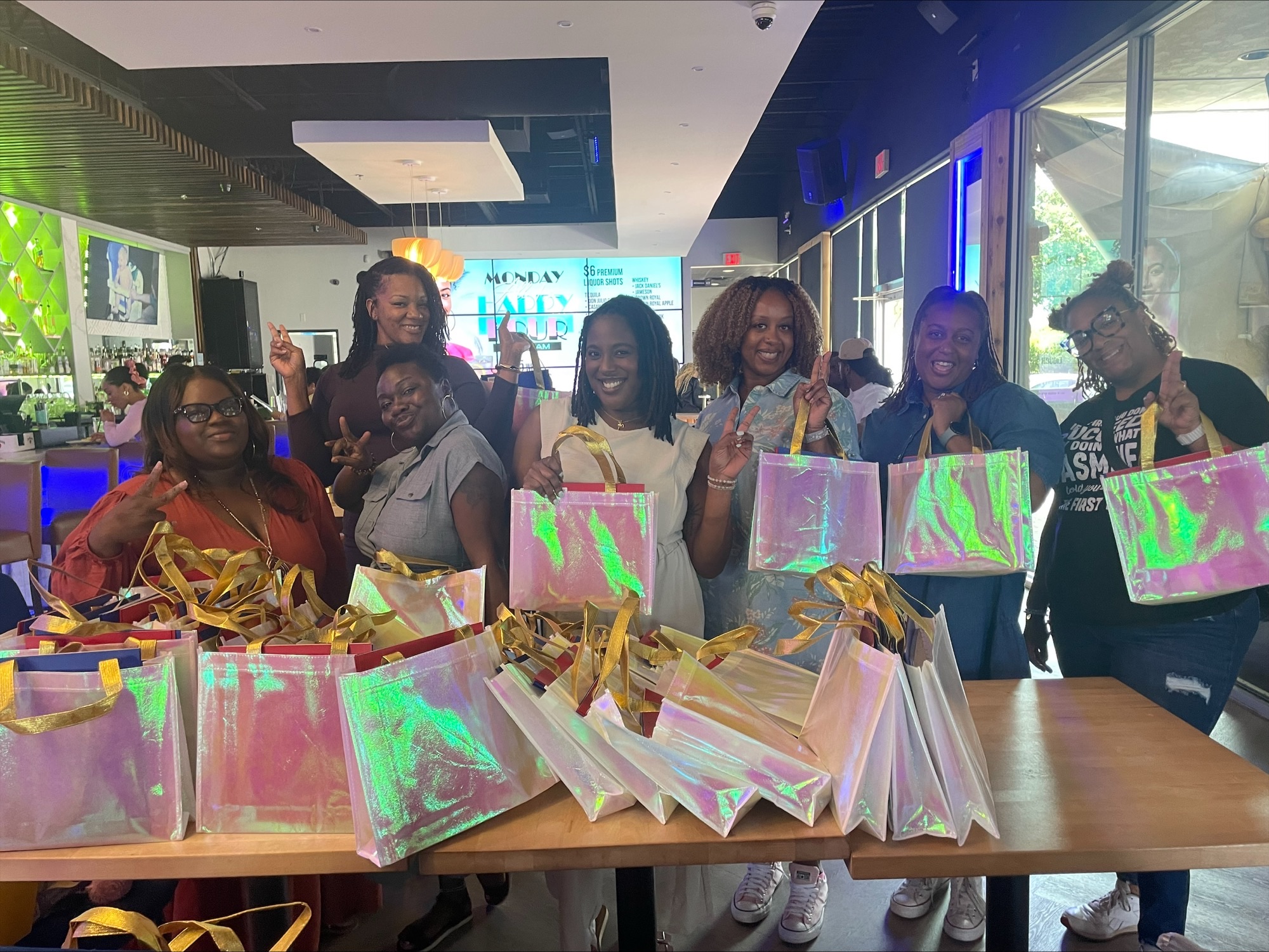 Group of women standing behind a table with colorful bags, smiling indoors with screens and windows in background.