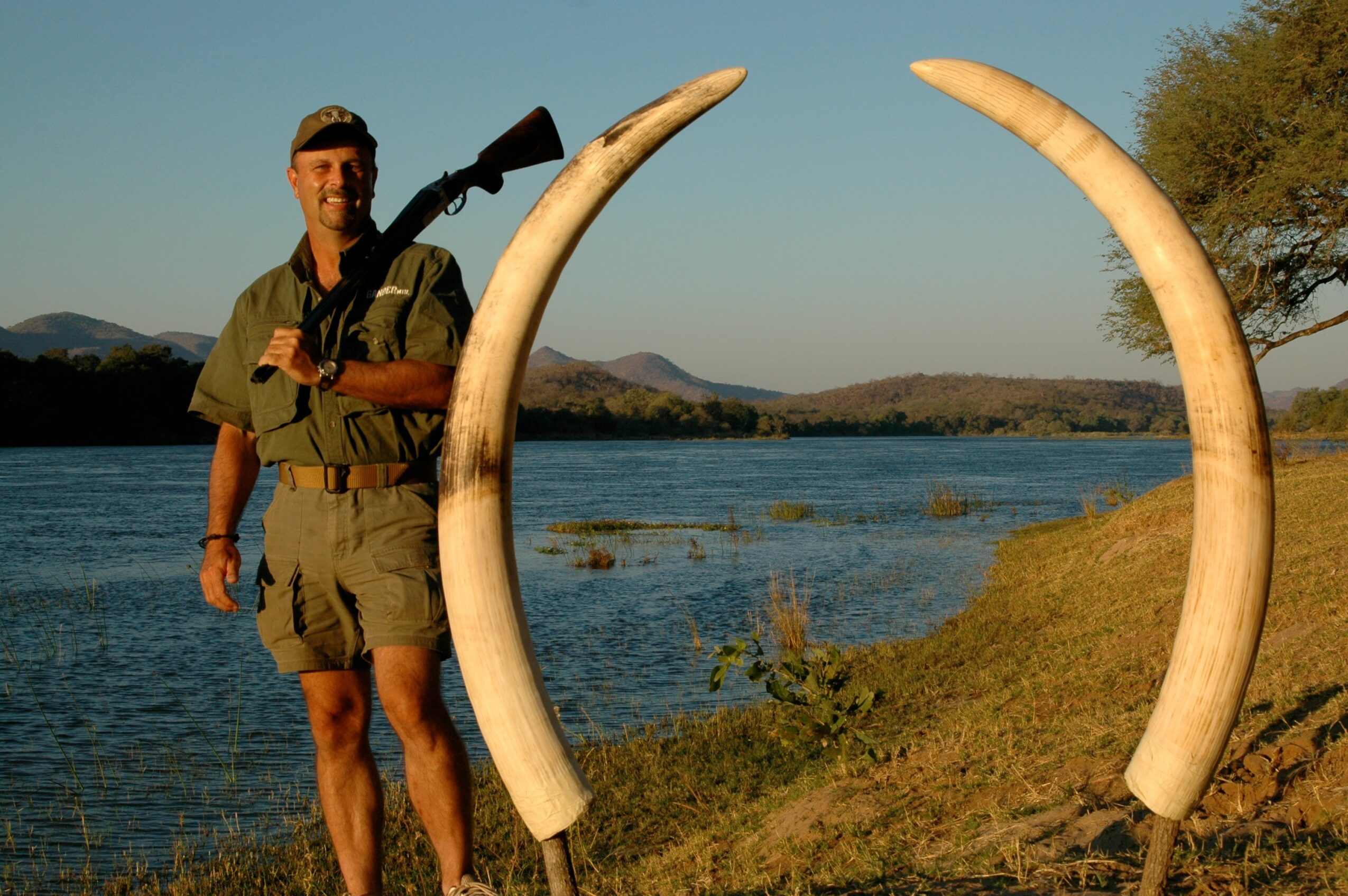 Person standing outdoors near water with large curved tusks, landscape with trees and mountains in background.
