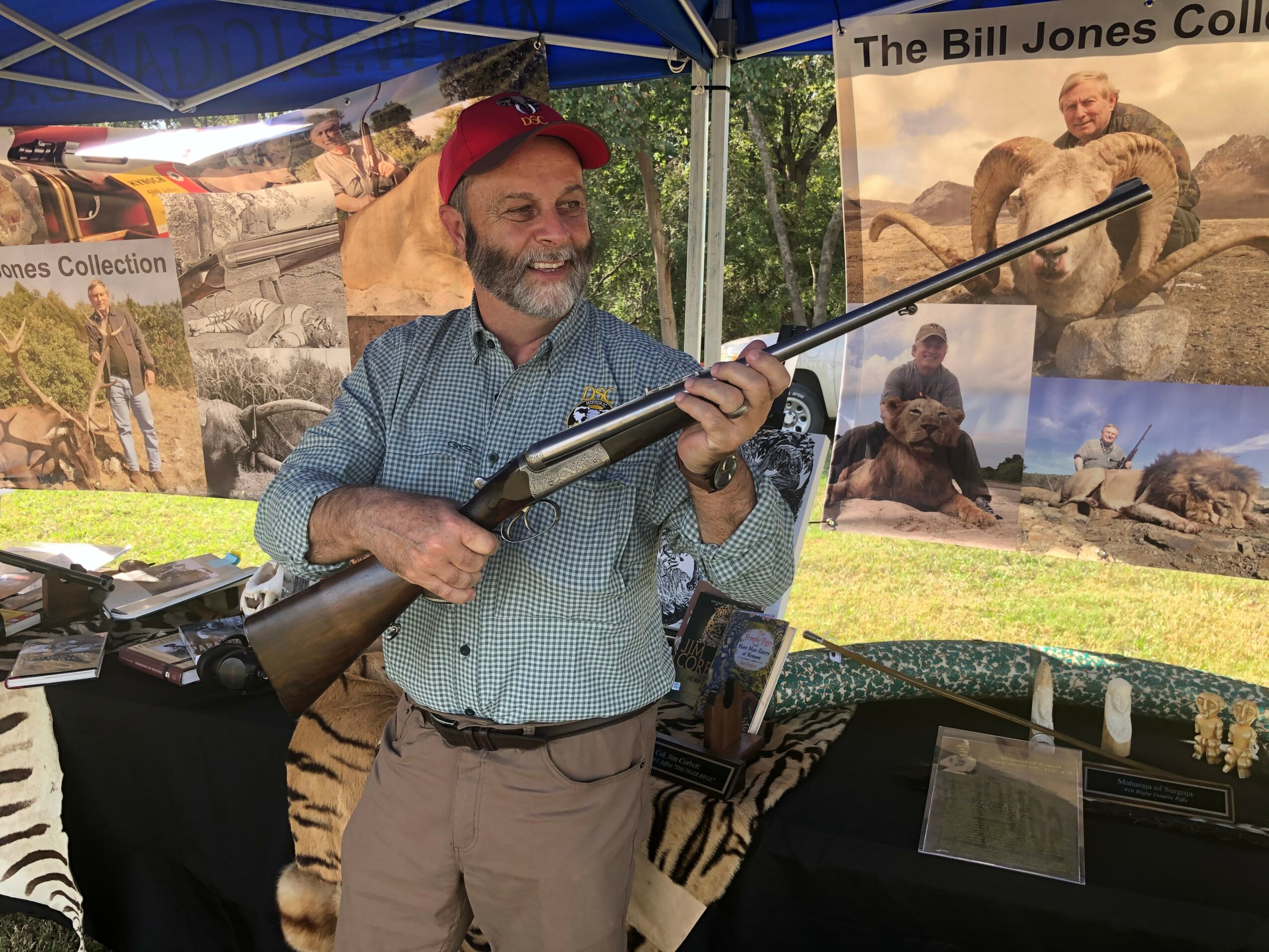 Man with a beard and red cap holding a rifle at an outdoor event, with posters and banners in the background.