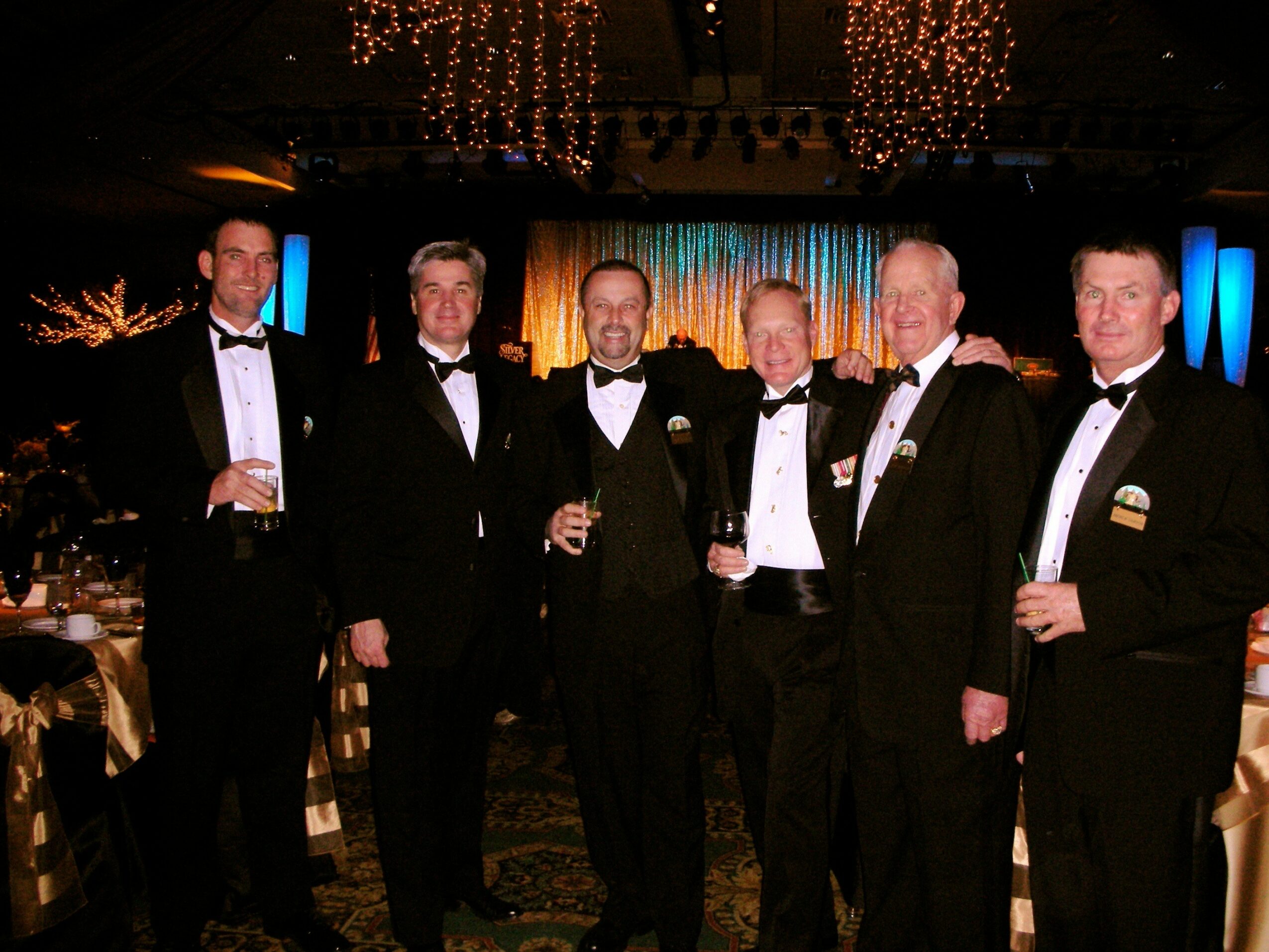 Six men in tuxedos pose together at a formal event, holding drinks, with a decorated stage in the background.