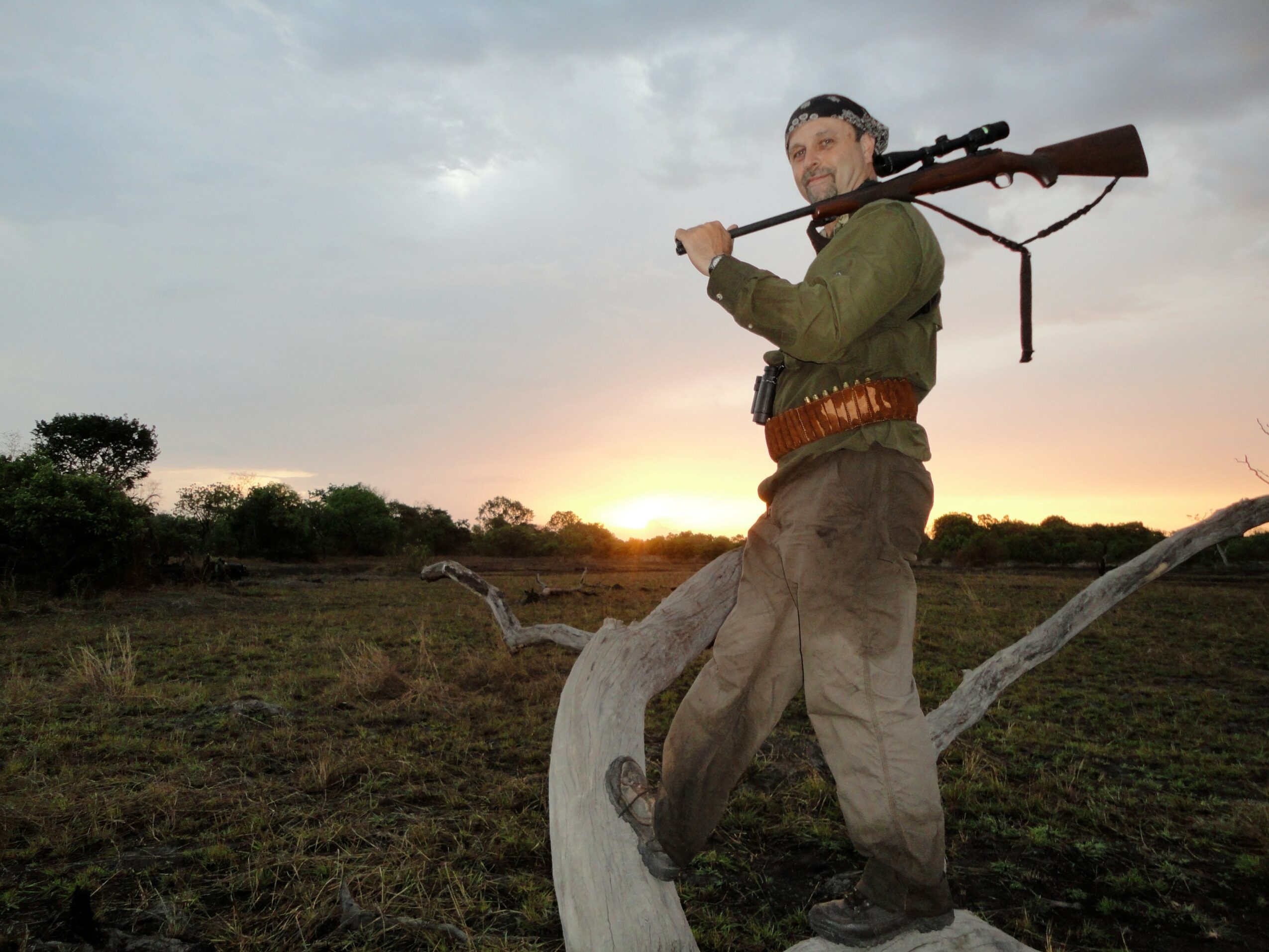 Man standing outdoors holding a rifle over his shoulder during sunset, with a tree branch in the foreground.