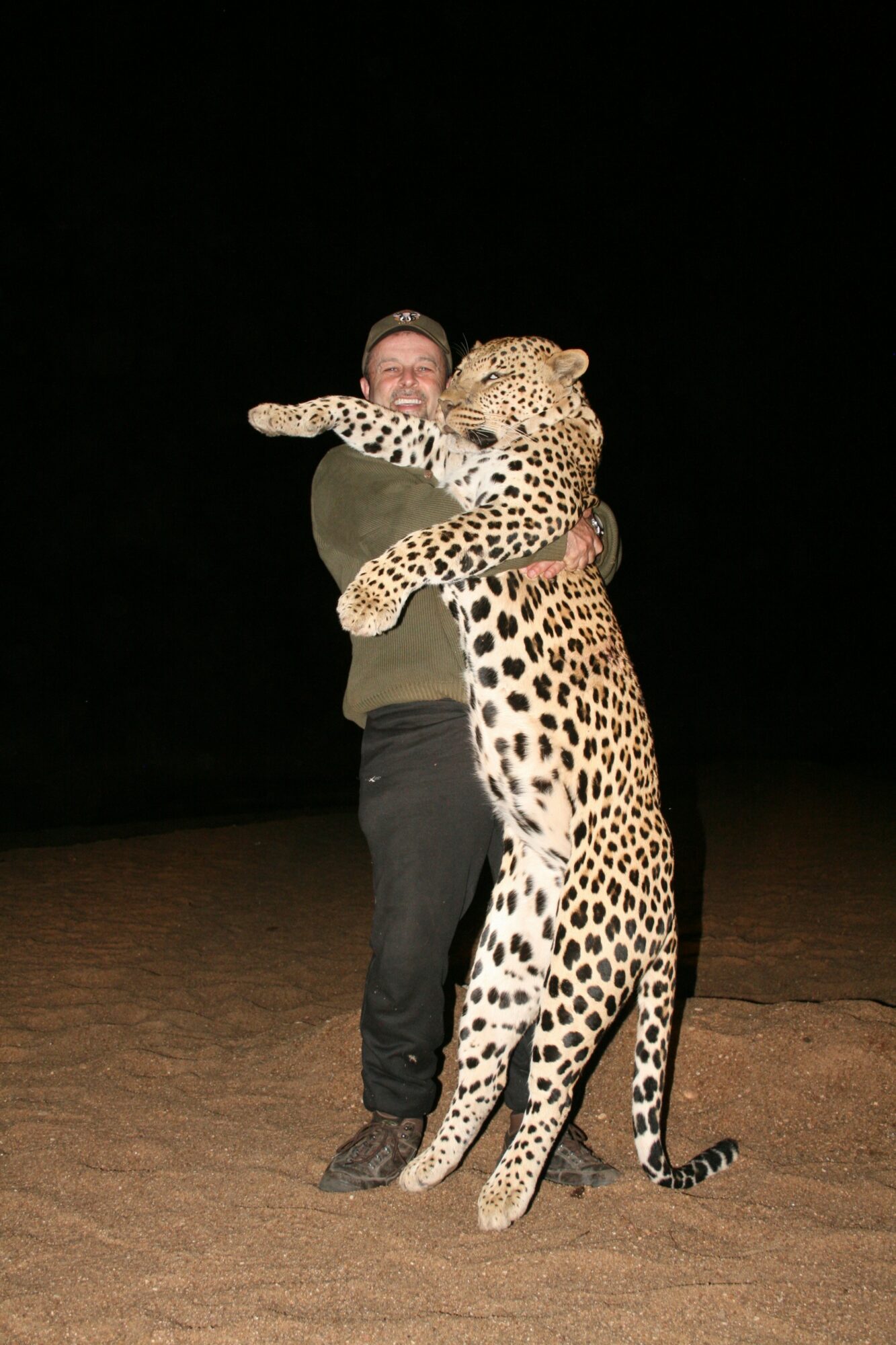 Person hugging a large cheetah at night, with a dark background, ground visible, and the cheetah's spots visible.