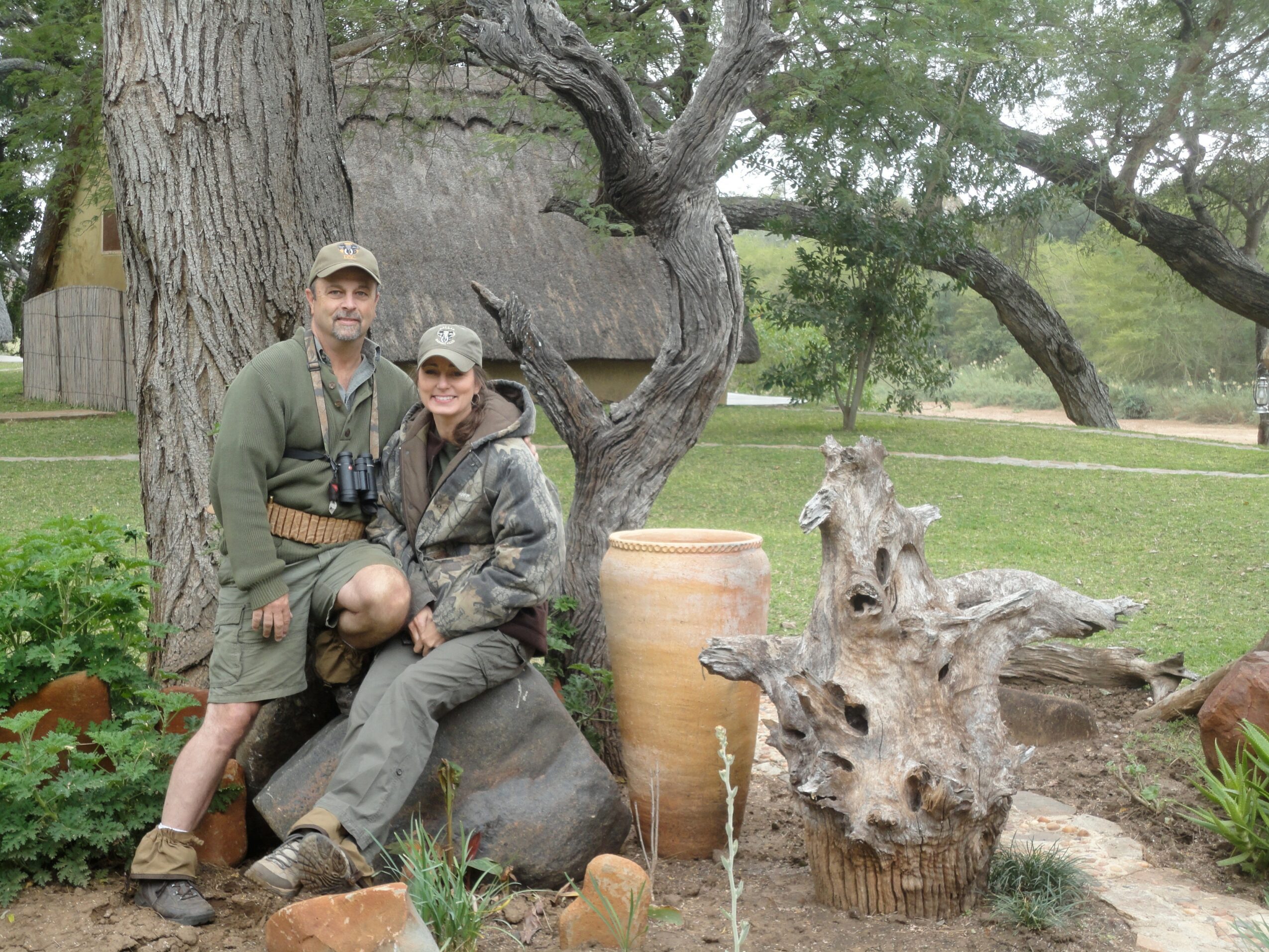 Two people sitting on a large rock outdoors with trees and a large clay pot nearby.