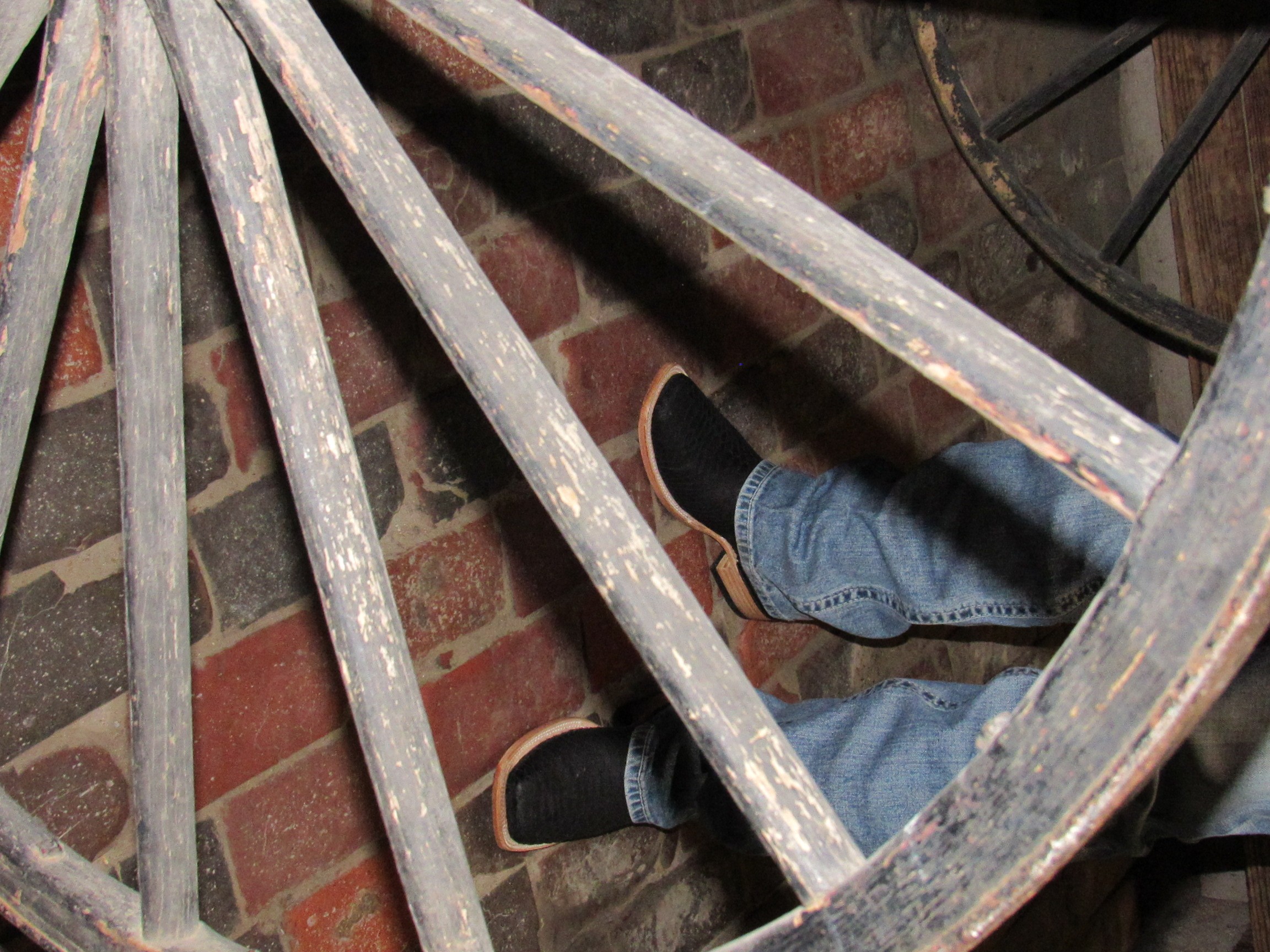 Person's legs and feet in jeans and black shoes resting on a rusty metal spiral staircase against a brick wall.