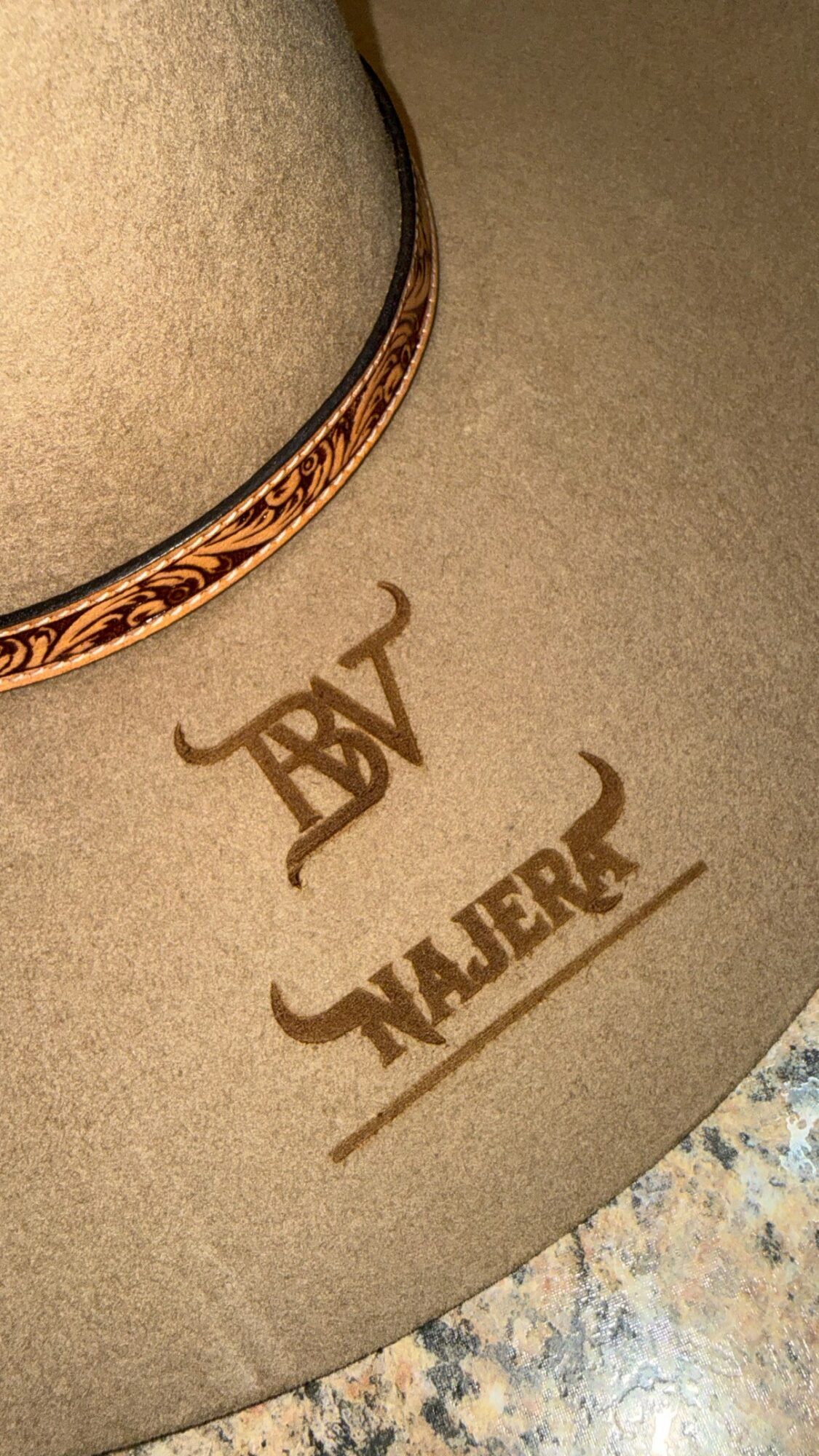 Close-up of a beige hat with decorative band, text reading 'ABT' and 'NAJER' underneath, on a speckled surface.