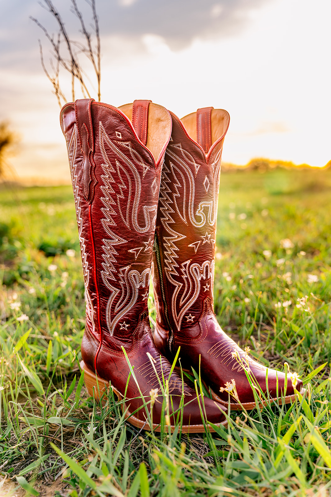 Pair of tall red cowboy boots with decorative stitching, standing on grass in an outdoor field during sunset.