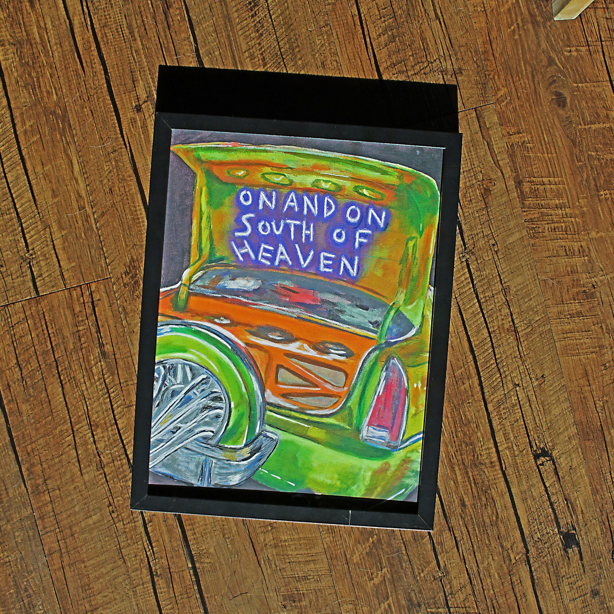 Colorful painting of a vehicle with a sign reading 'ON AND ON SOUTH OF HEAVEN' inside a black frame, on a wooden floor.