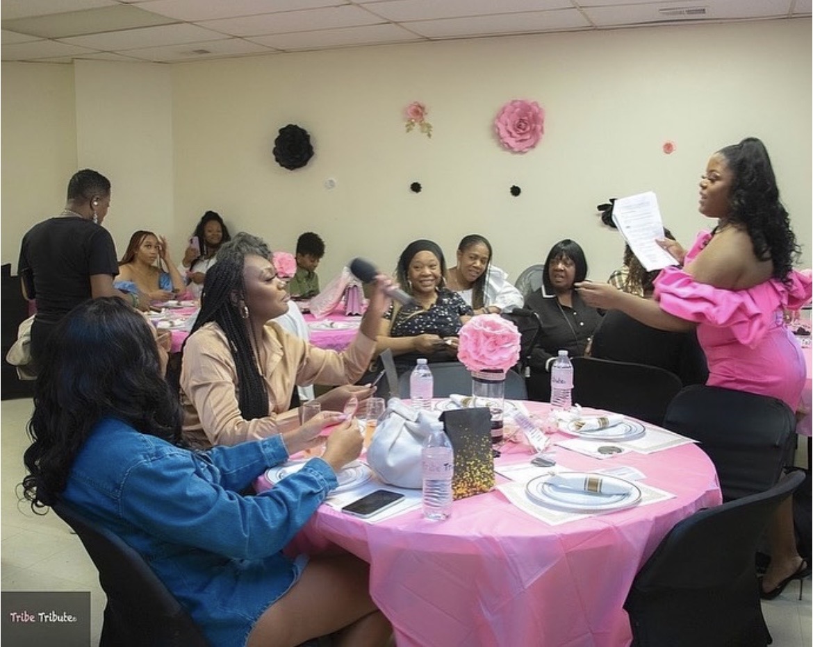 Group of women sitting at a table with pink tablecloth, listening to a woman standing and reading from paper, decorated with pink and black flowers