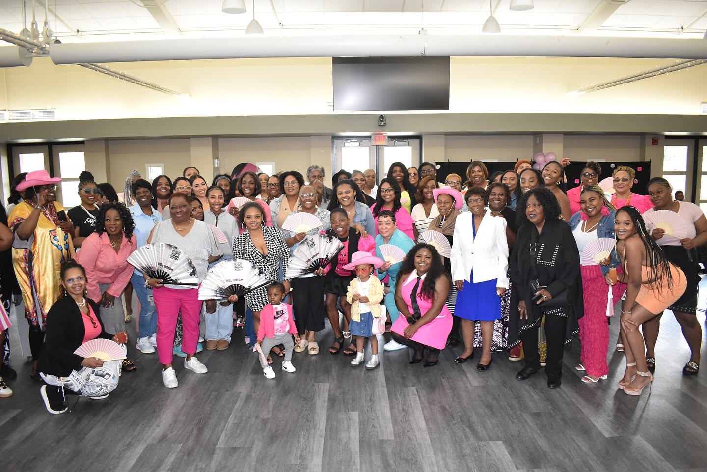 Group of women, children, and a few men gathered indoors, some wearing pink hats, in a spacious room with a large screen.