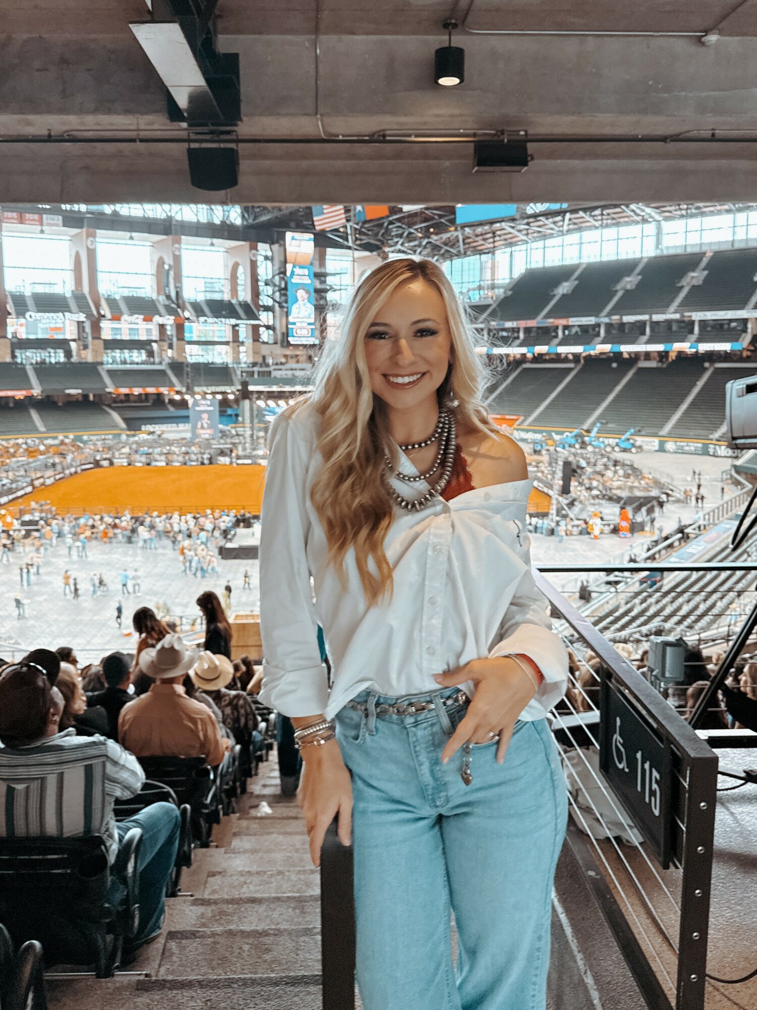 Smiling woman with long blonde hair in a white off-shoulder top and jeans at a stadium, with spectators and field in background.